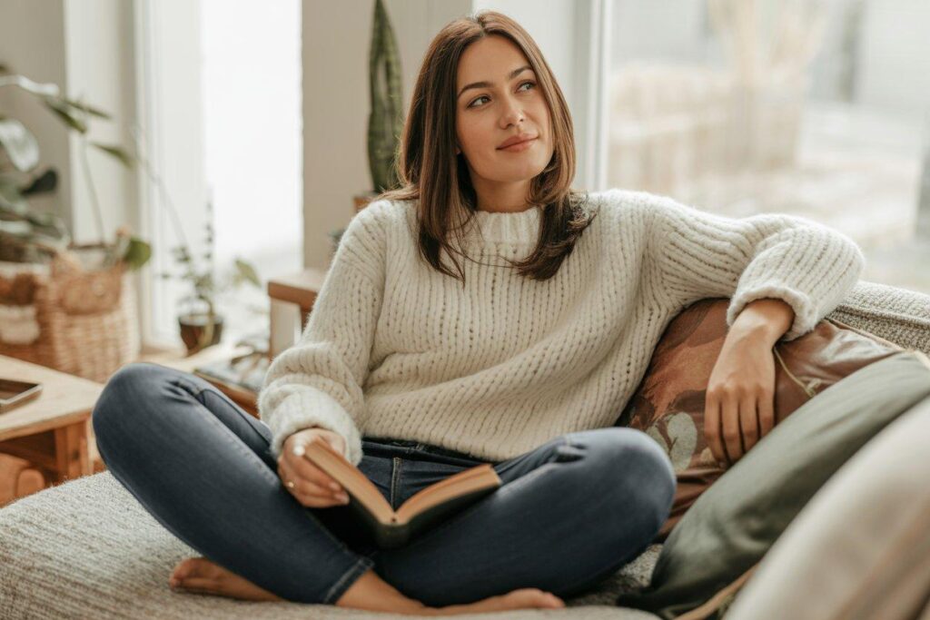 Woman in cream chunky sweater and dark skinny jeans reading book on living room sofa