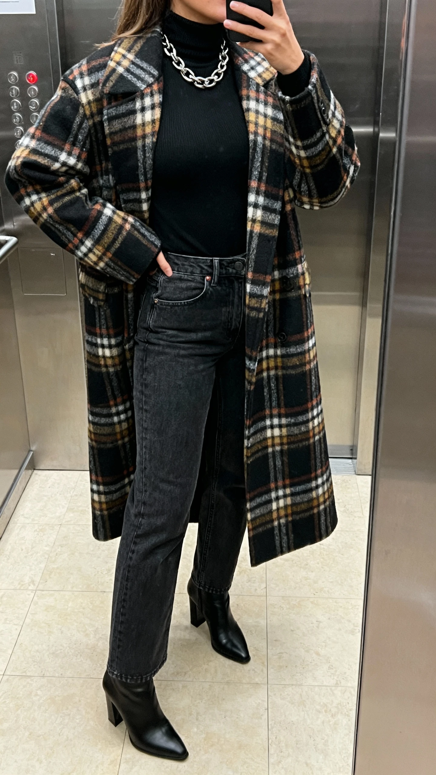 iPhone back-view selfie in elevator mirror of a woman wearing an all-black base: fitted turtleneck and high-rise straight jeans with black heeled boots, topped with a bold plaid statement coat, chunky chain necklace peeking at collar; face not visible, indoor lighting, natural iPhone photo quality.