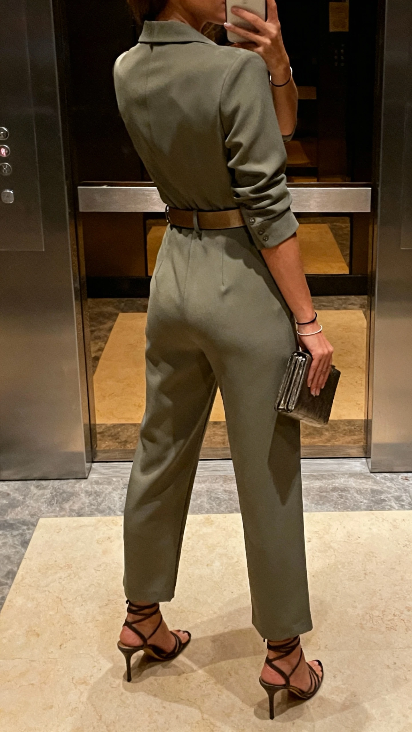 iPhone back-view selfie of a woman wearing an elevated tailored jumpsuit, waist-defining belt, strappy heels, clutch, thin bracelet; face not visible, elevator mirror, evening indoor lighting, natural iPhone quality.