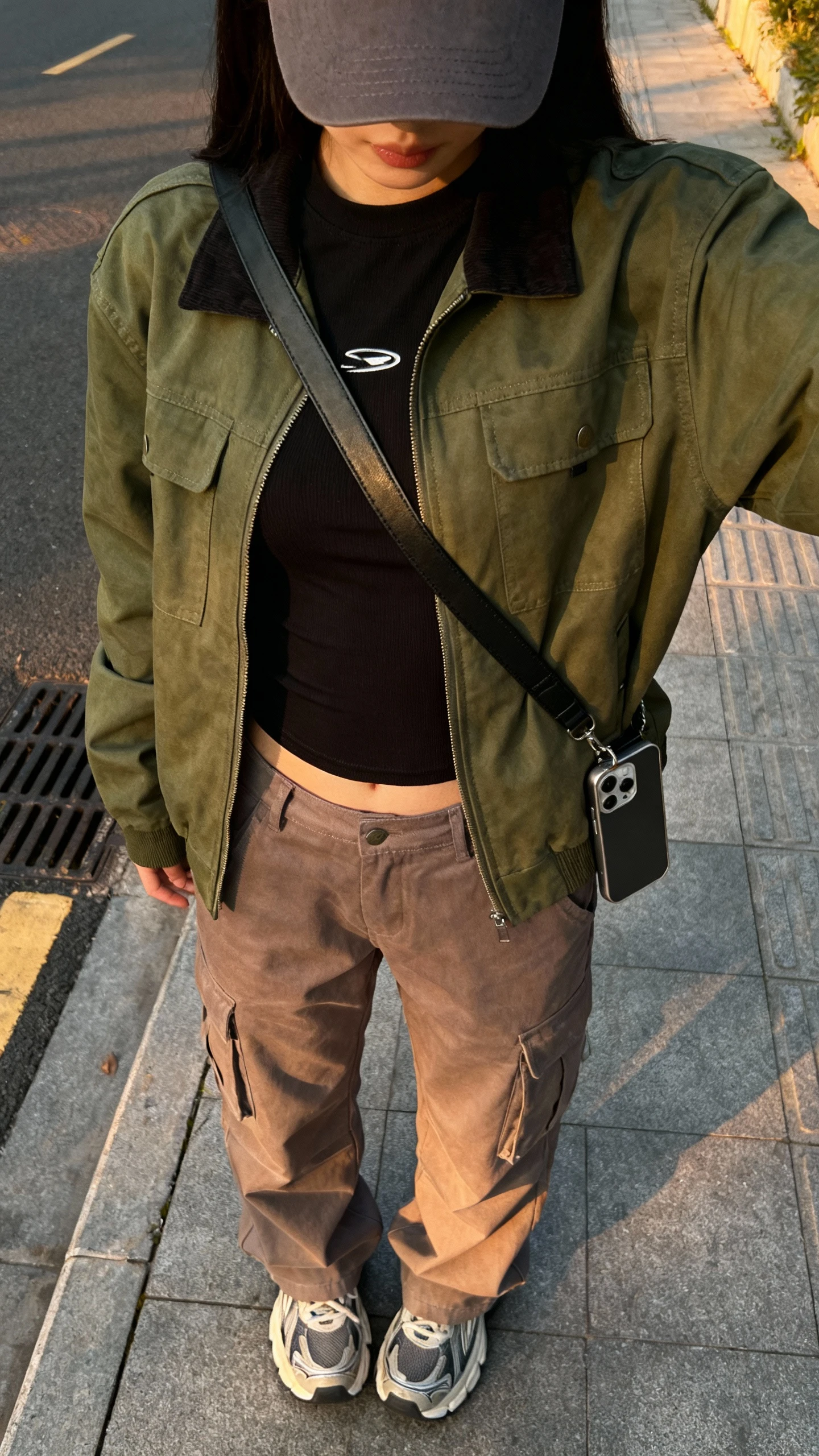 iPhone back-view street selfie of a woman wearing an olive utility jacket over a black fitted tee, taupe cargo pants, chunky sneakers, baseball cap, crossbody phone bag, face not visible, sidewalk setting in soft evening light, natural iPhone photo quality.