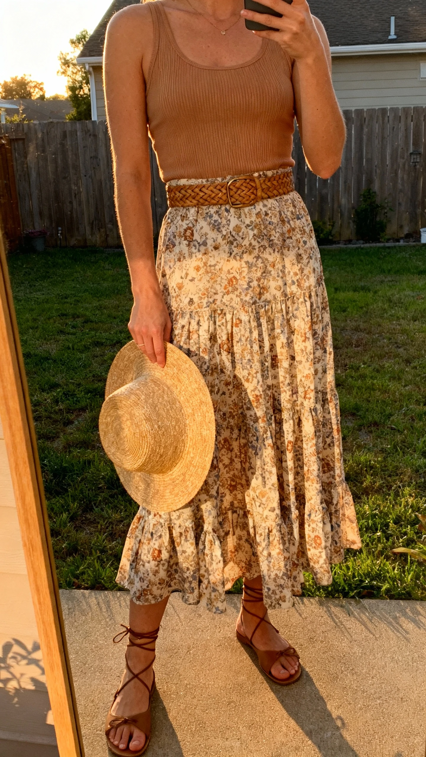 iPhone backyard mirror selfie of a woman wearing a flowy printed maxi skirt with a tan ribbed tank, woven belt, flat lace-up sandals, and a straw hat in hand; face not visible, golden-hour light, casual iPhone photo.