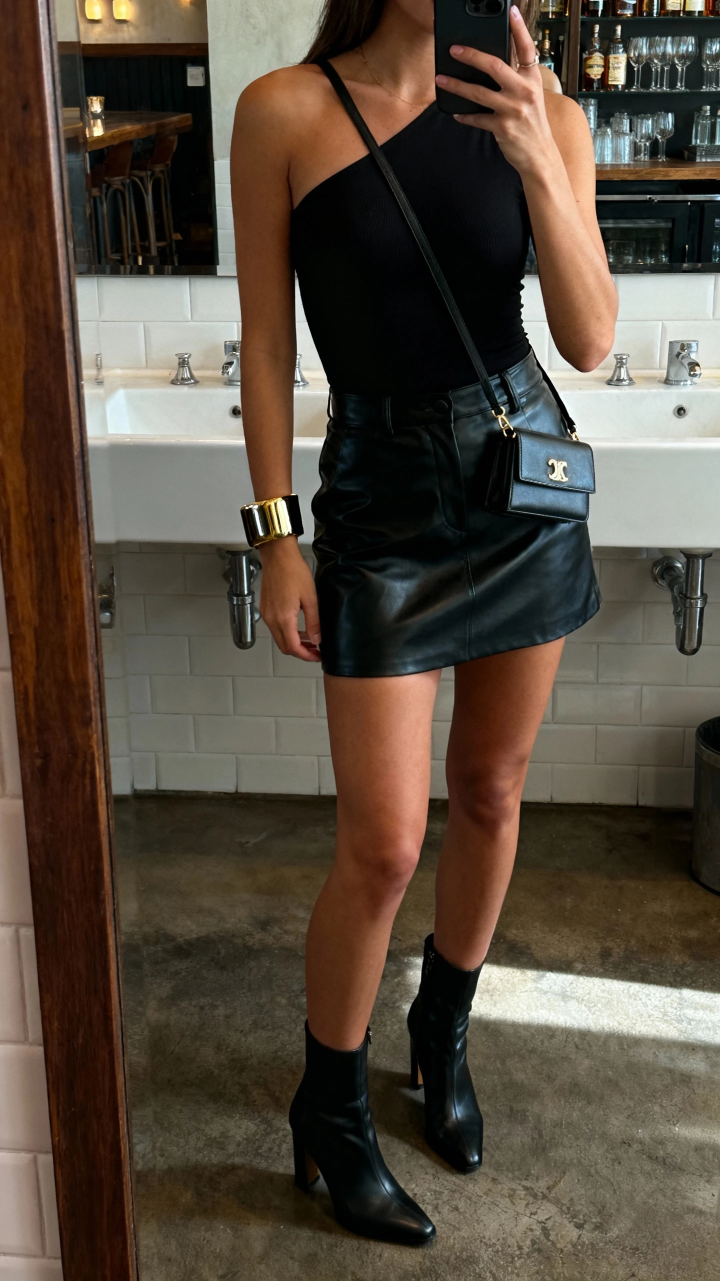 iPhone bar-bathroom mirror selfie of a woman wearing a leather-skirt night-out formula: black one-shoulder bodysuit, high-waisted black leather mini skirt, heeled ankle boots, mini crossbody, bold cuff bracelet, face not visible, moody indoor lighting, natural iPhone aesthetic.