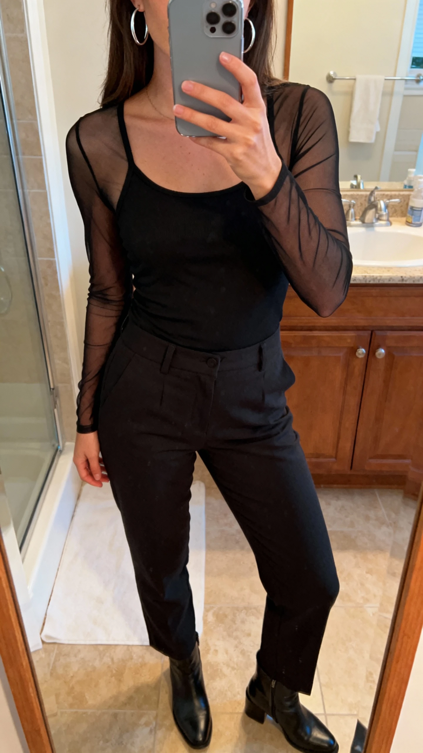 iPhone bathroom mirror selfie of a woman layering a sheer long-sleeve black mesh top over a black tank, with straight-leg trousers, ankle boots, and silver hoops; face not visible, warm indoor lighting, natural iPhone photo.