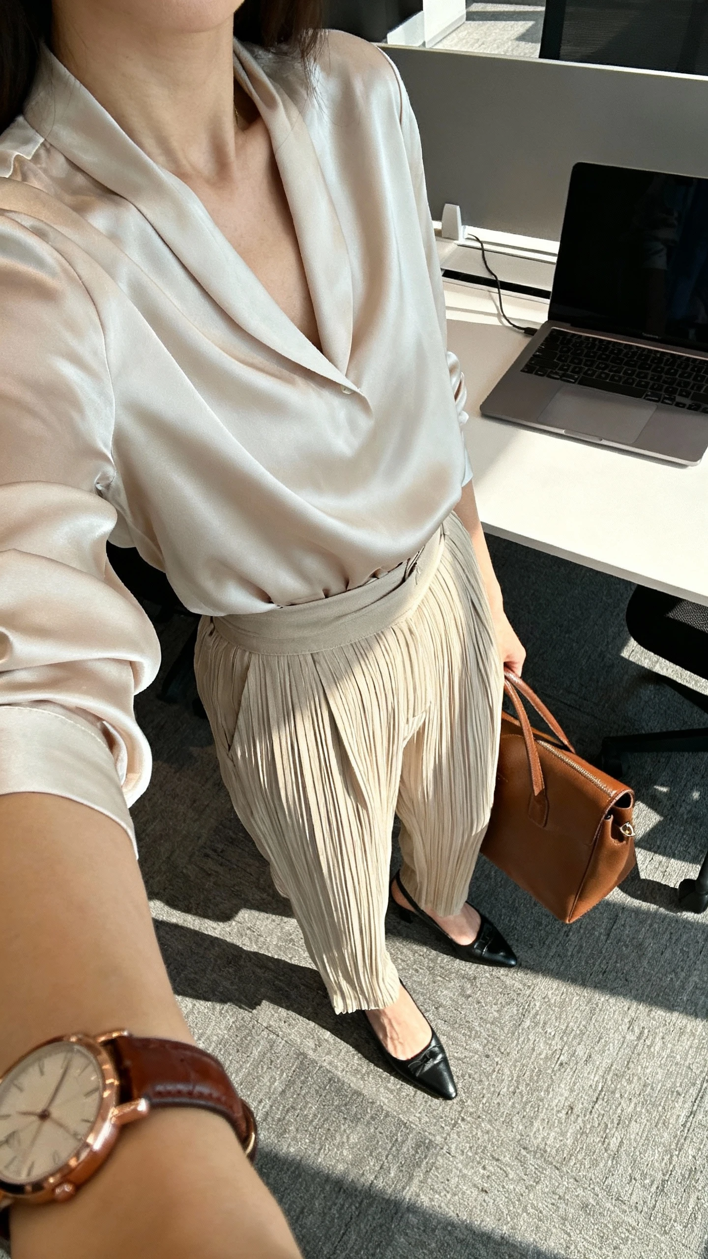 iPhone desk-side selfie cropped at shoulders of a woman in pleated trousers with a silk blouse tucked in, classic pumps, leather watch, laptop bag; face not visible, modern office with morning light, natural iPhone photo.
