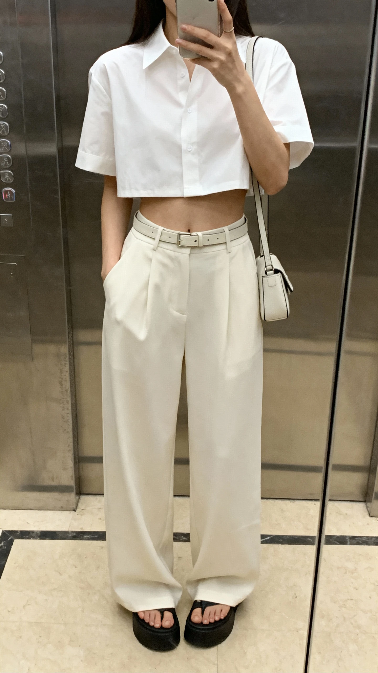 iPhone elevator mirror selfie of a woman in a cropped boxy blouse in crisp white, paired with high-rise wide-leg trousers, minimalist belt, platform sandals, small shoulder bag, face not visible, indoor lighting, natural iPhone photo quality