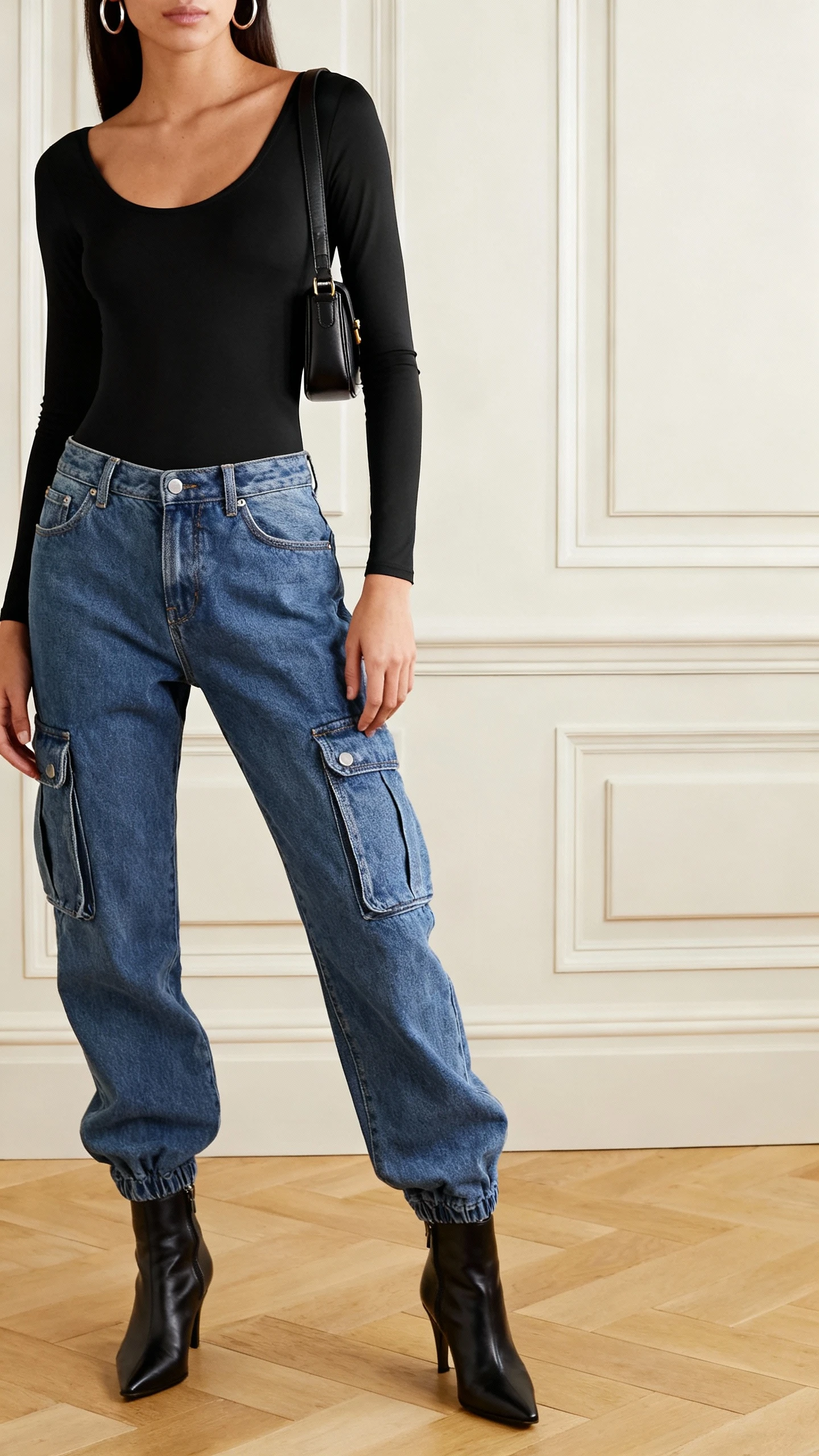iPhone elevator mirror selfie of a woman wearing denim cargo pants with a sleek black bodysuit, sharp ankle boots, mini shoulder bag, hoop earrings; face not visible, cropped at neck, indoor lighting, natural iPhone photo quality.