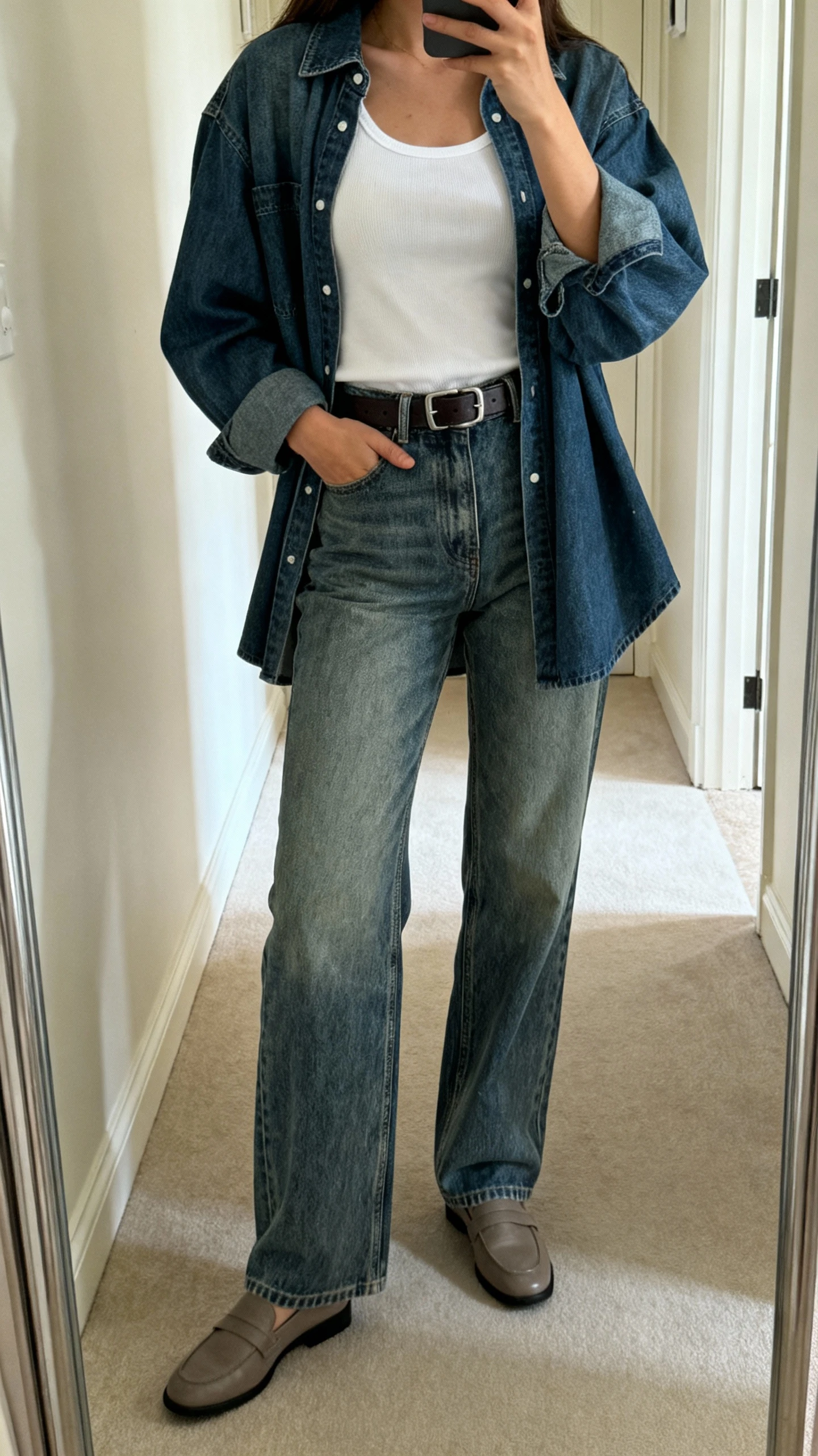 iPhone full-length mirror selfie of a woman wearing a white tank tucked into high-rise medium-wash denim jeans with a matching oversized denim shirt jacket, belt, and loafers; face not visible, hallway mirror, natural daylight, iPhone quality.