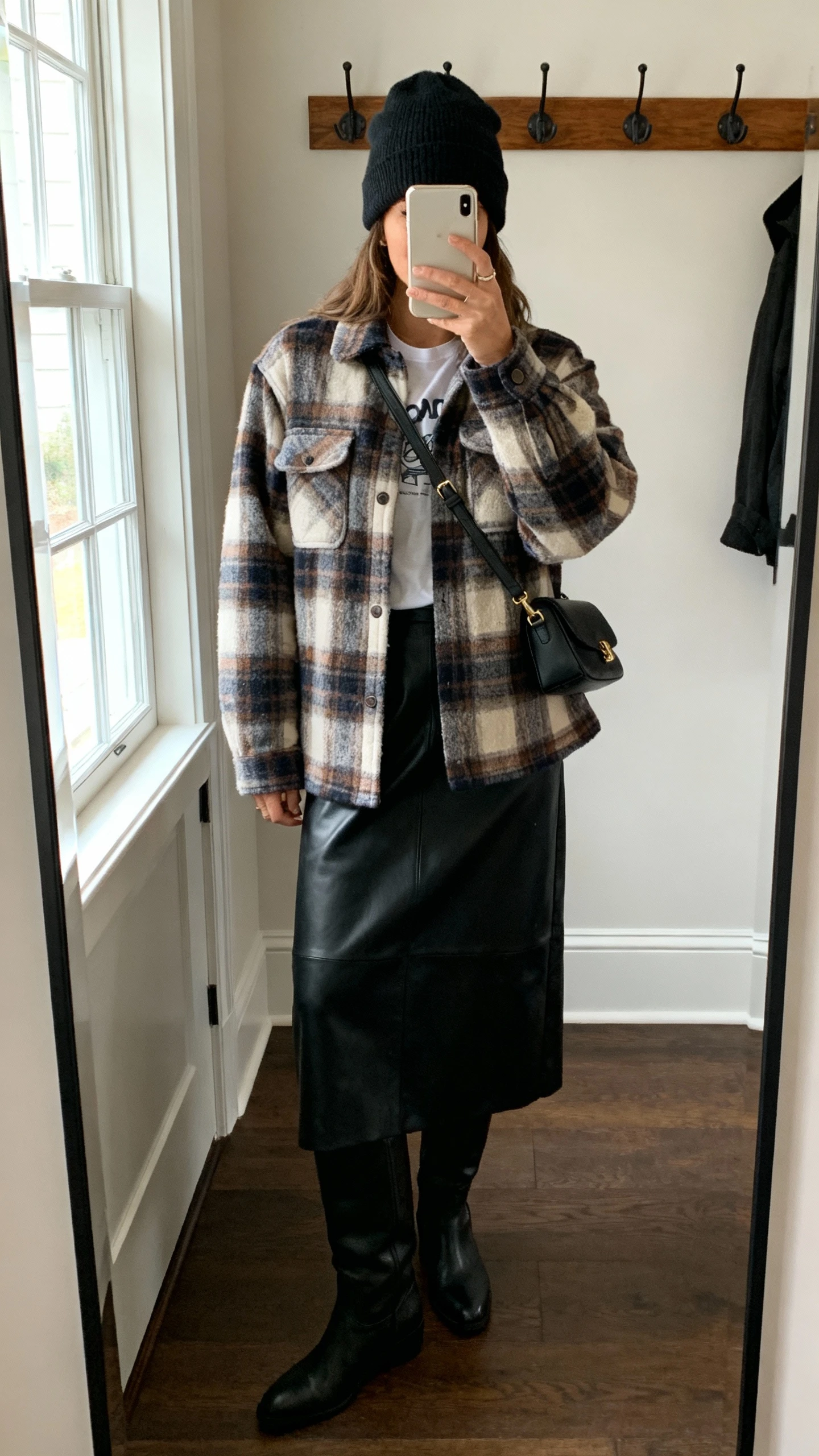 iPhone hallway mirror selfie of a woman wearing a cozy plaid shacket over a tee with a black leather midi skirt and tall boots, beanie, crossbody, face not visible, cropped at neck, entryway mirror with coat hooks, natural window light, casual iPhone photo