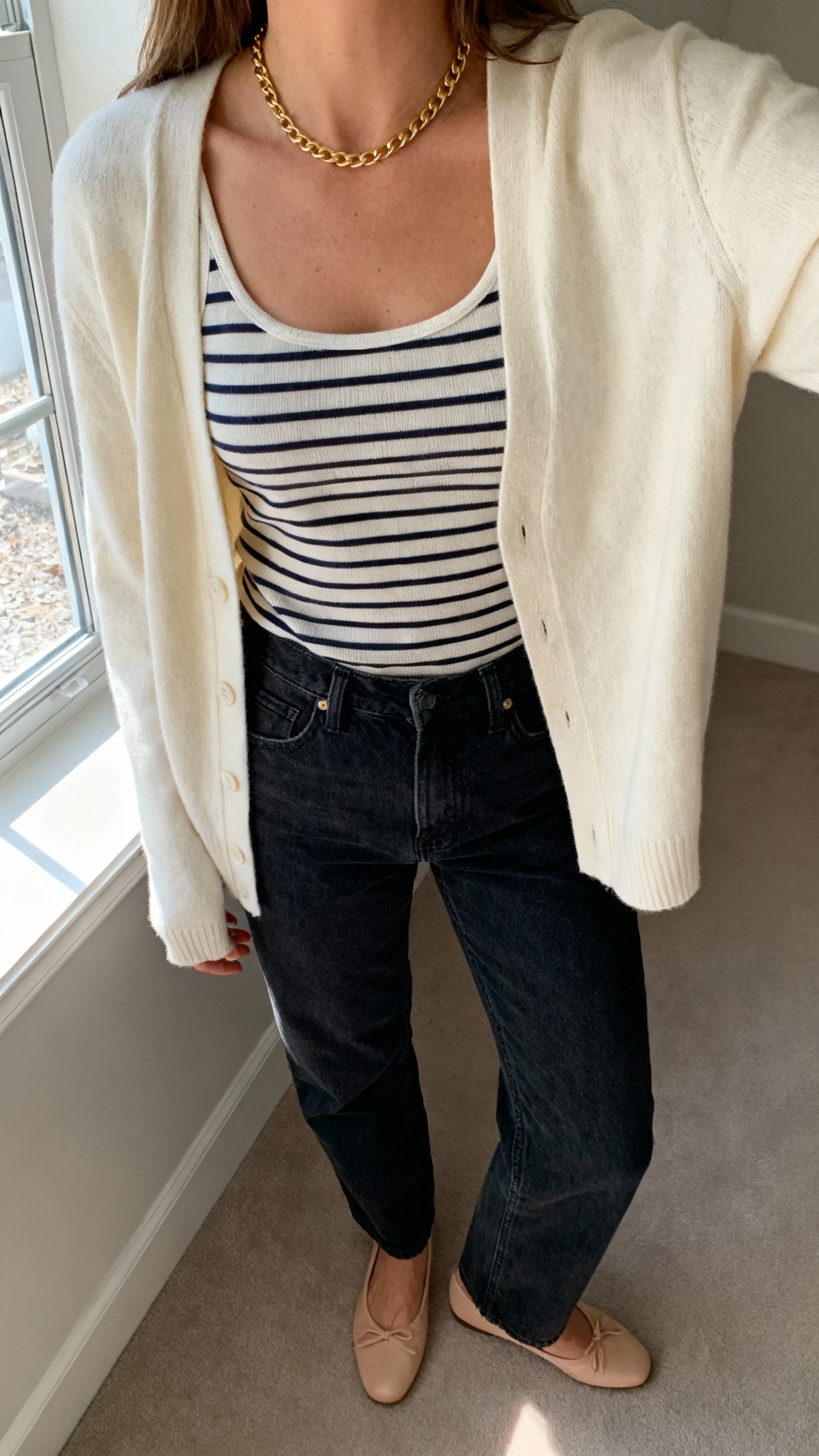 iPhone living-room mirror selfie of a woman in a cream cardigan worn open over a striped tank, dark-wash straight jeans, ballet flats, and a simple gold chain; face not visible, cropped at neck, natural window light, iPhone photo quality.