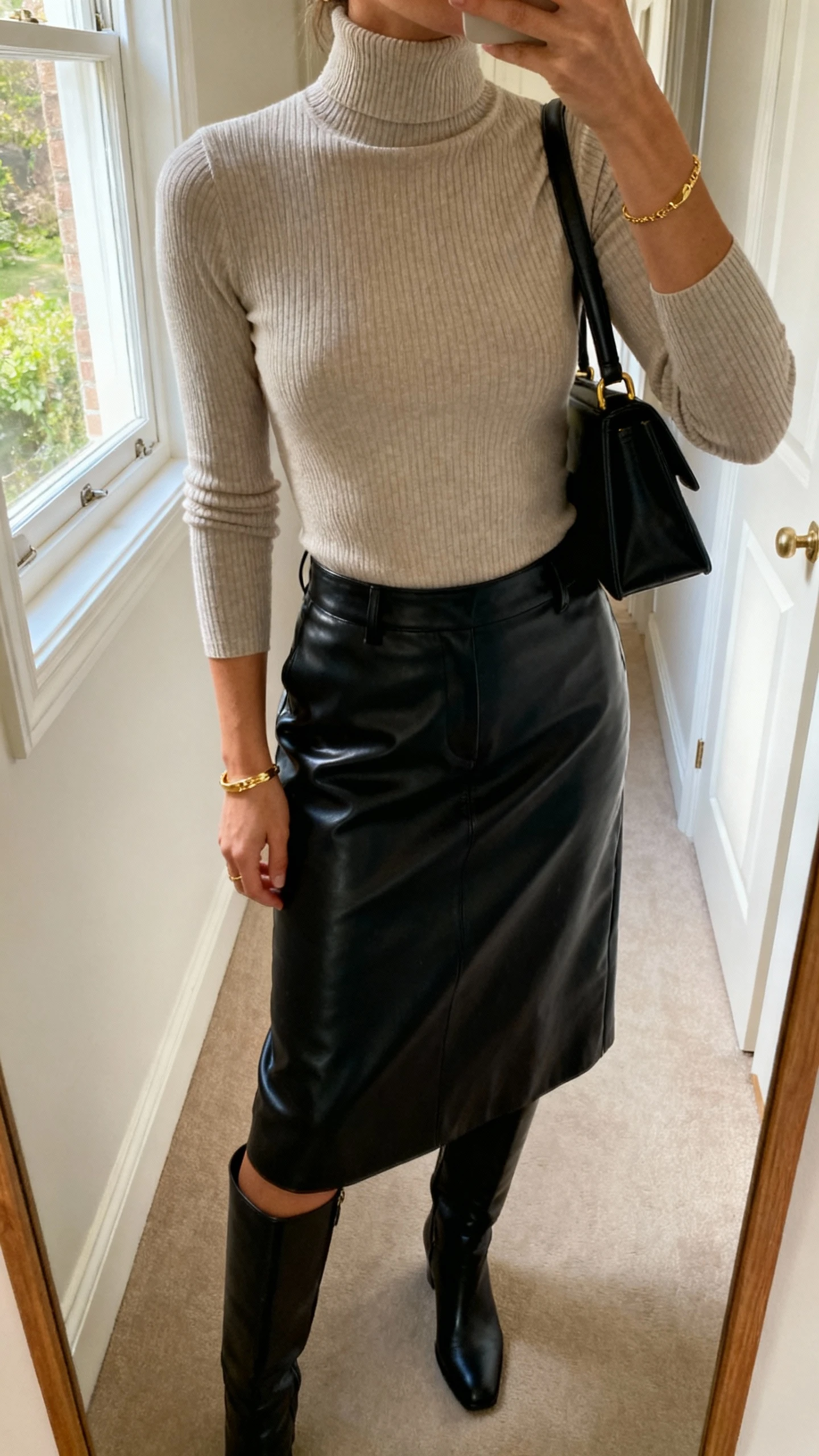 iPhone mirror selfie of a woman in a fitted ribbed turtleneck paired with a black leather midi skirt and knee-high boots, minimal gold jewelry, top-handle bag, face not visible, cropped at neck, hallway mirror, morning window light, natural iPhone quality
