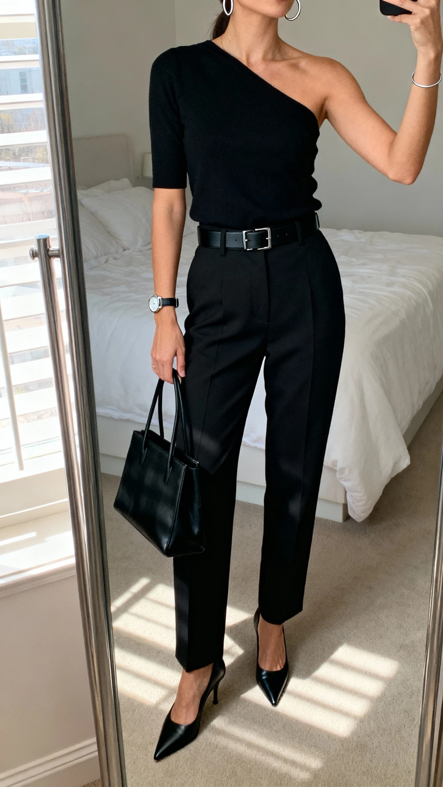 iPhone mirror selfie of a woman in a minimalist monochrome look: black one-shoulder top tucked into high-waisted black tailored trousers, sleek black belt, pointed black heels, structured black tote, thin silver hoops and watch, face not visible, cropped at neck, clean bedroom mirror with morning window light, natural iPhone photo quality.
