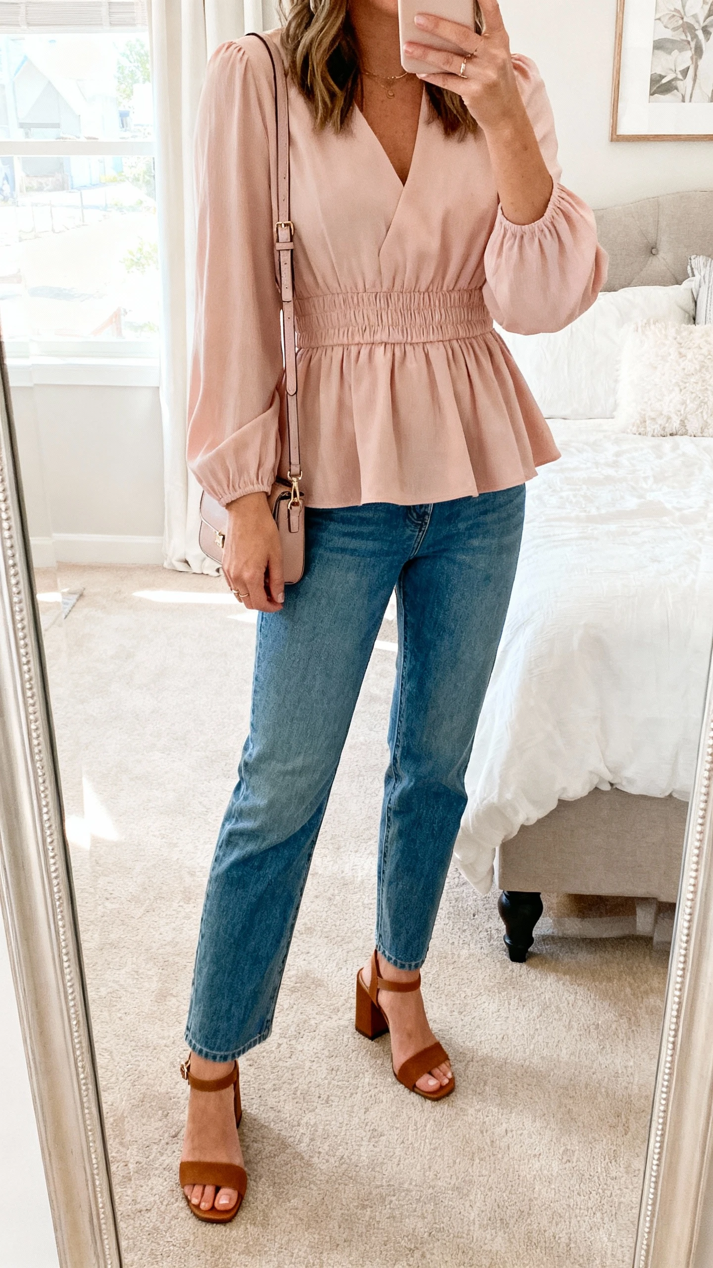 iPhone mirror selfie of a woman in a peplum blouse in soft blush with a comfortable smocked waist, paired with straight-leg blue jeans, block-heel sandals, small crossbody bag, face not visible, cropped at shoulders, bedroom mirror, bright natural daylight, natural iPhone aesthetic