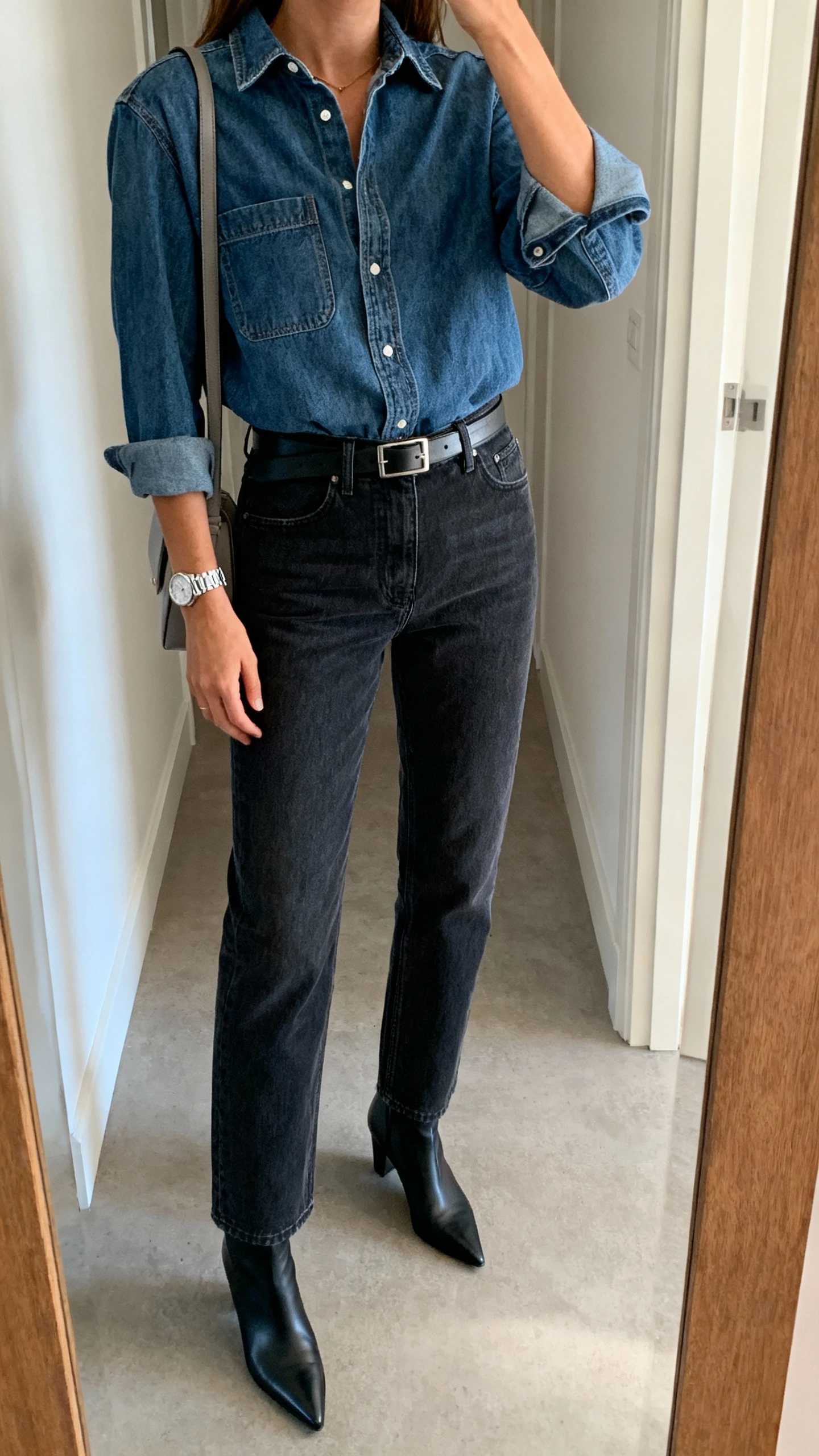 iPhone mirror selfie of a woman in elevated denim-on-denim: structured medium-wash denim shirt tucked into straight-leg dark-wash jeans, pointed ankle boots, slim leather belt, minimalist shoulder bag, silver watch, face not visible, cropped at shoulders, hallway mirror with morning light, natural iPhone photo quality.