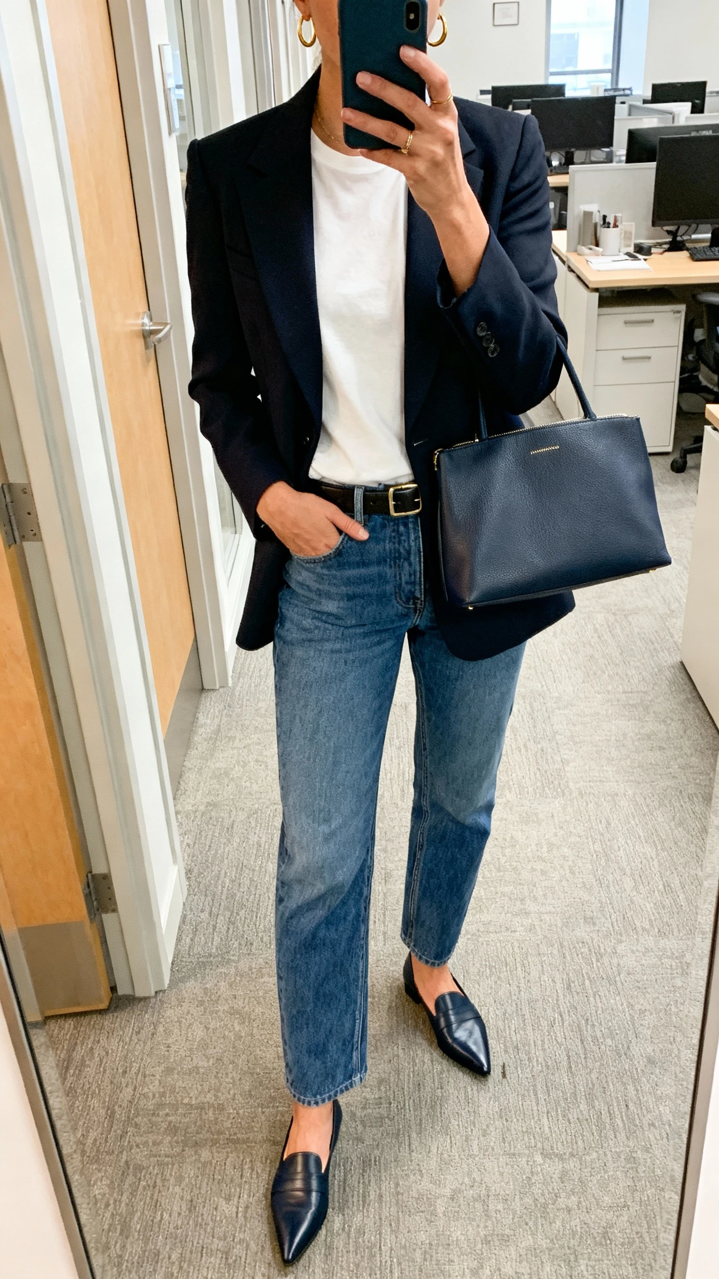iPhone mirror selfie of a woman wearing a crisp tailored blazer over a white tee, straight-leg blue jeans, pointed loafers, slim belt, structured tote, minimal gold hoops; office hallway mirror, cropped at neck, face not visible, natural morning light, natural iPhone photo quality.