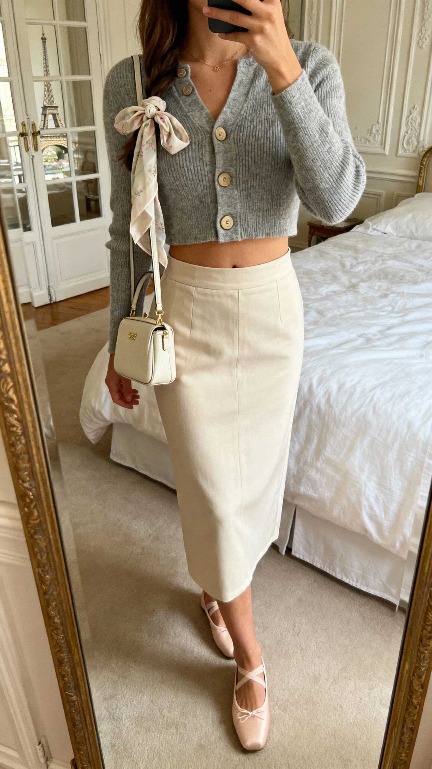 iPhone mirror selfie of a woman wearing a cropped cardigan buttoned as a top with a high-waisted skirt, ballet flats, delicate scarf tied to a mini bag; face not visible, Parisian-style bedroom mirror, soft daylight, natural iPhone quality.