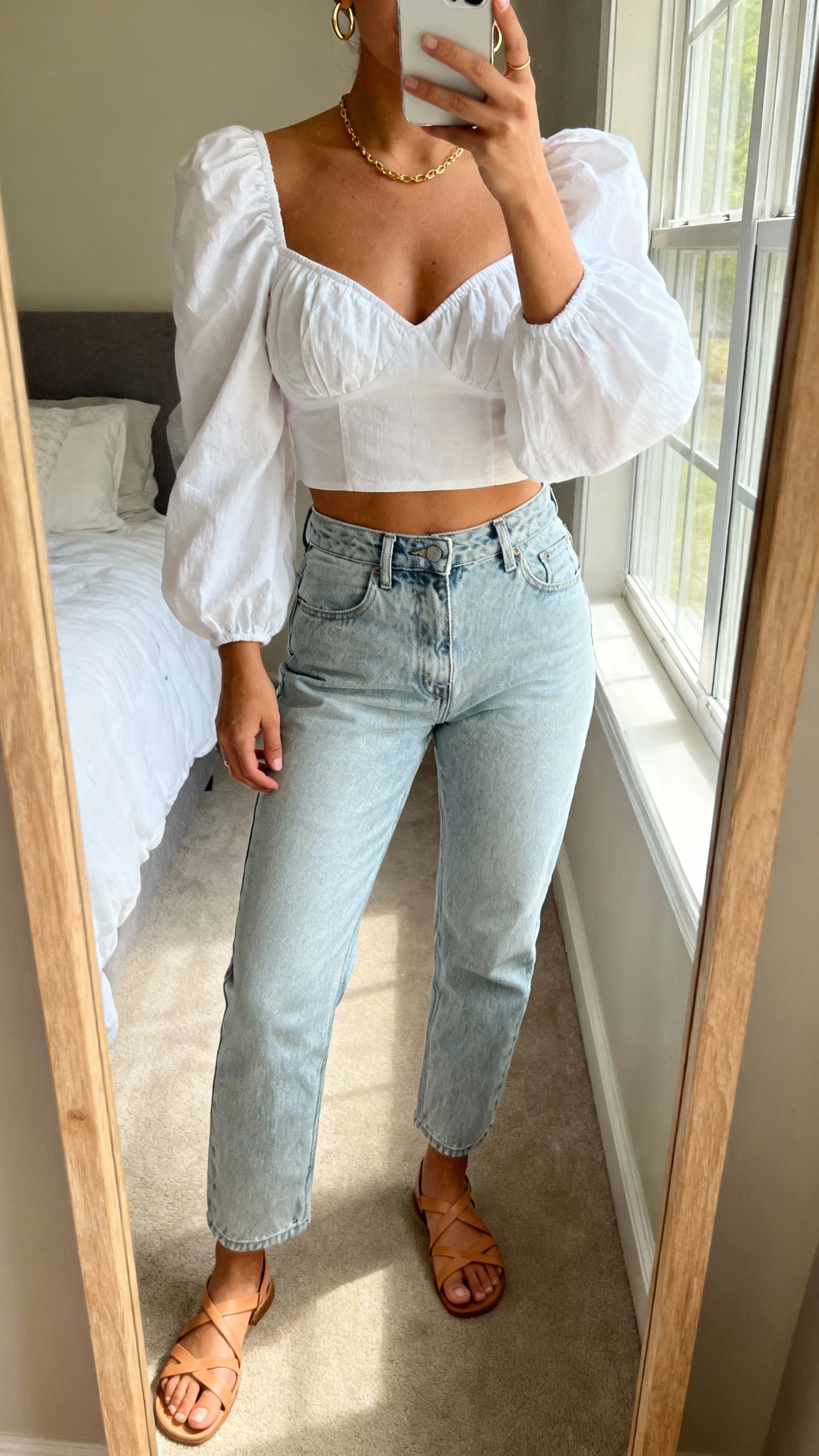 iPhone mirror selfie of a woman wearing a romantic white puff-sleeve blouse with sweetheart neckline, high-waisted light-wash jeans, delicate gold necklace, dainty hoop earrings, tan strappy sandals, face not visible, cropped at neck, bedroom mirror with morning window light, natural iPhone photo quality