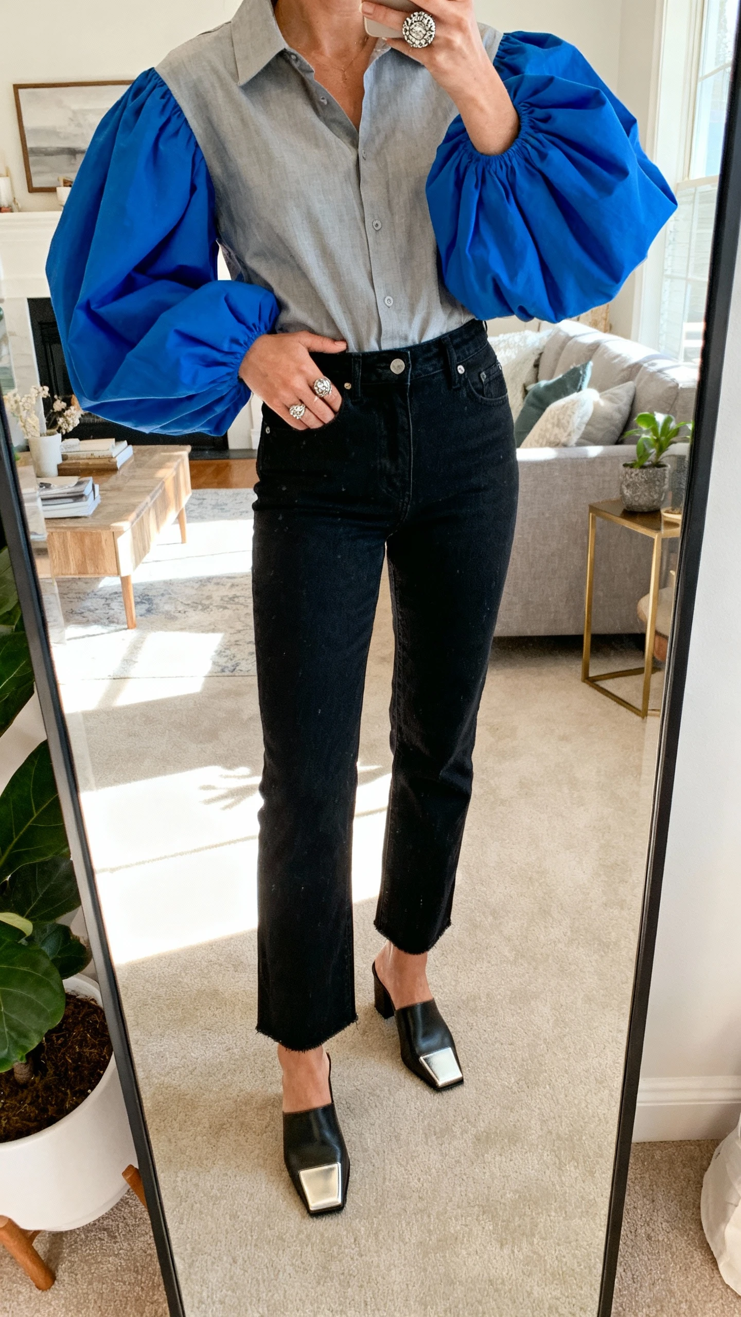iPhone mirror selfie of a woman wearing a statement sleeve blouse (bold balloon sleeves in cobalt), tucked into high-rise black jeans, square-toe mules, chunky ring, face not visible, full-length mirror in living room, bright afternoon light, casual iPhone photo