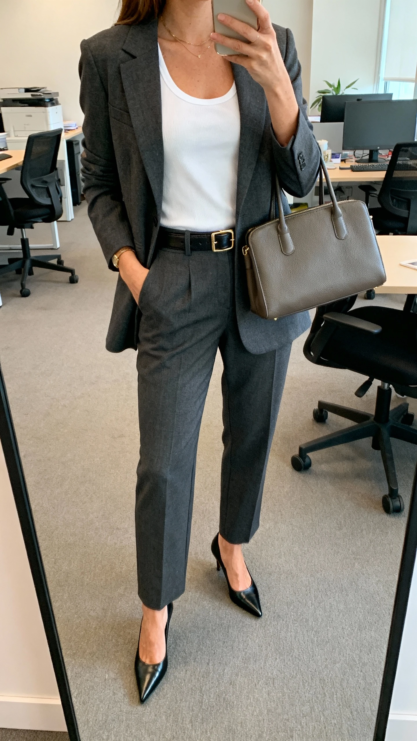 iPhone office mirror selfie of a woman wearing a charcoal blazer and matching trousers with a crisp white tank, pointed pumps, slim leather belt, and structured tote; face not visible, cropped at neck, morning office light, natural iPhone quality.