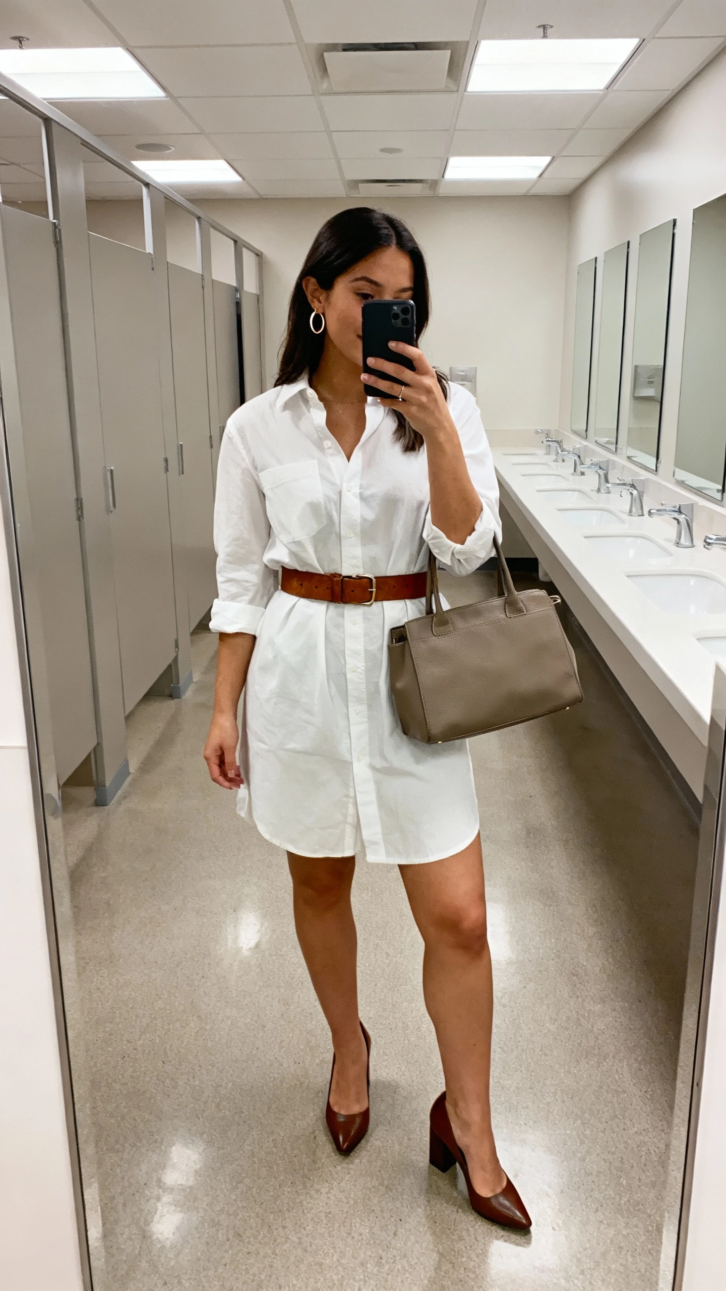 iPhone office restroom mirror selfie of a woman in a crisp shirt dress cinched with a leather belt, block-heel pumps, structured tote, subtle hoop earrings; face not visible, cropped at shoulders, overhead office lighting, natural iPhone quality.
