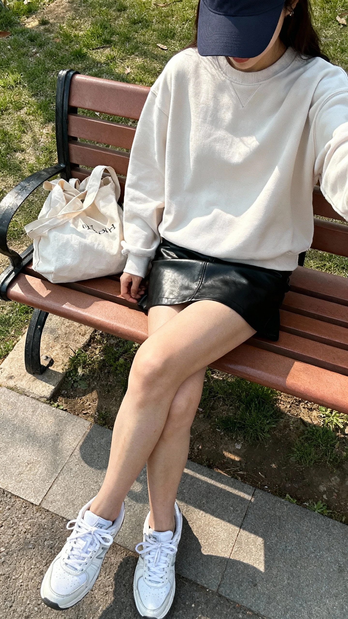 iPhone park bench selfie cropped at neck showing a woman in a relaxed crewneck sweatshirt with a black leather mini skirt and clean white sneakers, baseball cap, tote bag, face not visible, late-morning daylight, casual iPhone photo quality