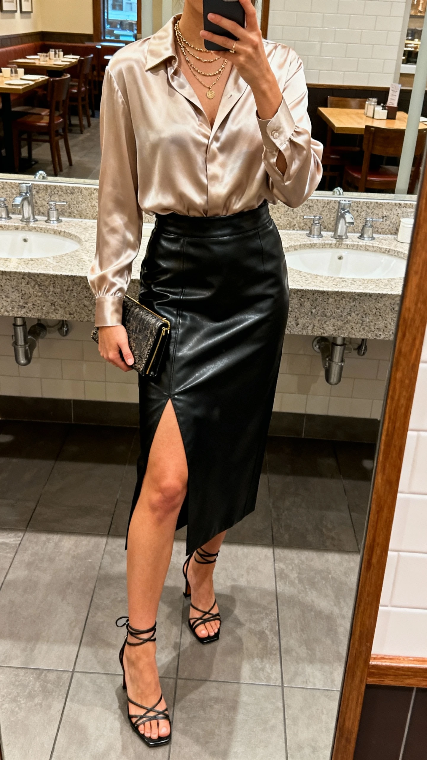 iPhone restaurant bathroom mirror selfie of a woman in a silky blouse tucked into a black leather slit midi skirt, strappy heels, clutch, layered necklaces, face not visible, cropped at shoulders, warm ambient lighting, natural iPhone aesthetic