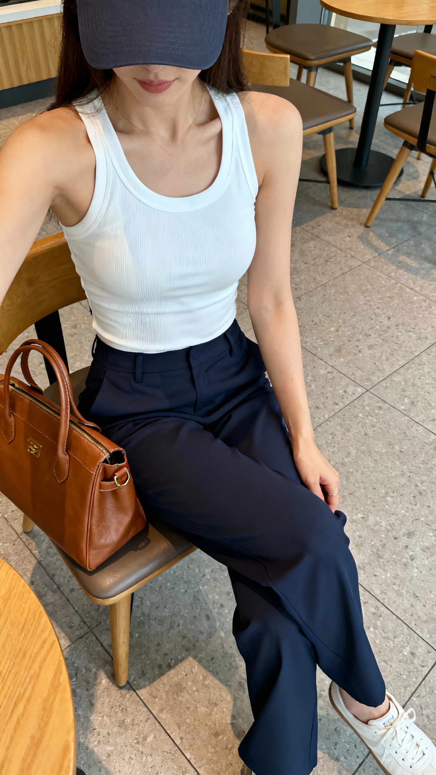 iPhone seated café selfie cropped at shoulders showing a woman in a white athletic tank, tailored navy trousers, slim white sneakers, baseball cap, and a leather tote on chair; face not visible, soft morning light, natural iPhone photo.