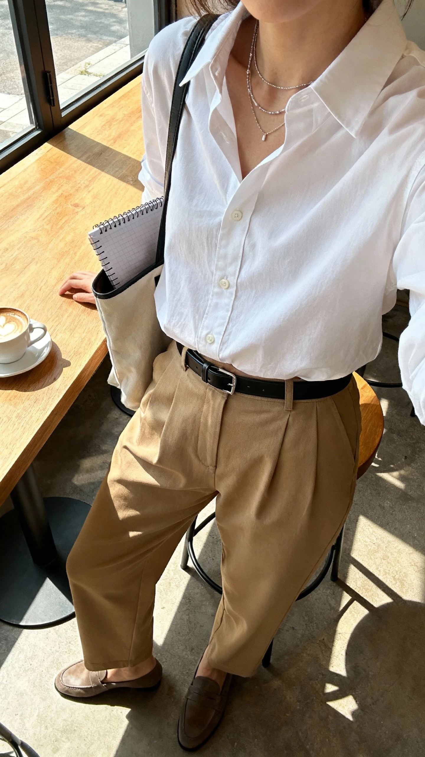 iPhone selfie cropped at shoulders showing a woman in a crisp white button-up half-tucked into relaxed tan trousers, loafers, slim black belt, delicate layered necklaces, tote with notebook peeking out, face not visible, coffee shop table setting with morning window light, natural iPhone photo quality.