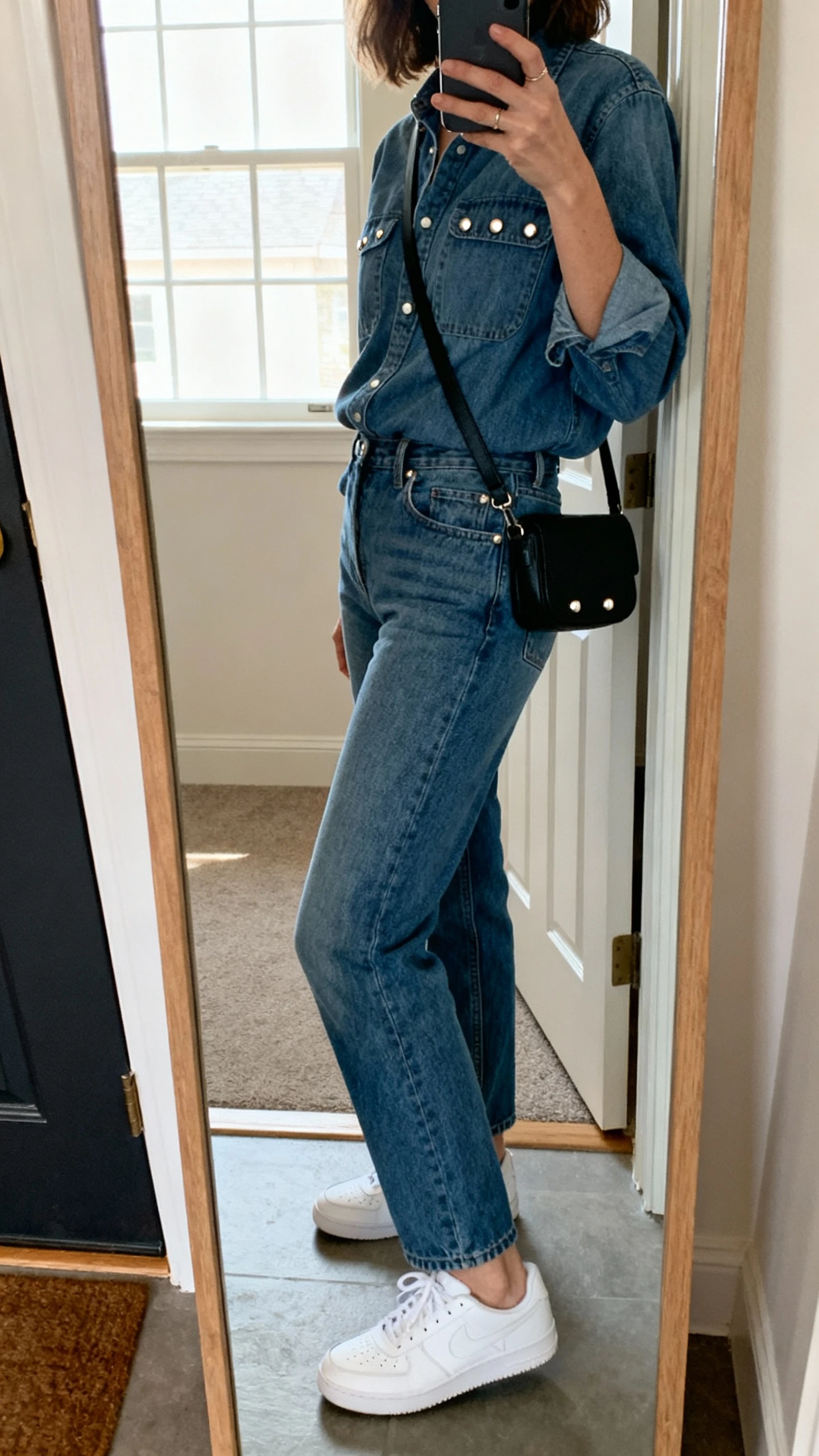 iPhone side-angle mirror selfie of a woman in a coordinated denim shirt and matching denim jeans (double denim set), clean white sneakers, simple black crossbody, understated studs; apartment entry mirror, face not visible, natural window light, casual iPhone aesthetic.