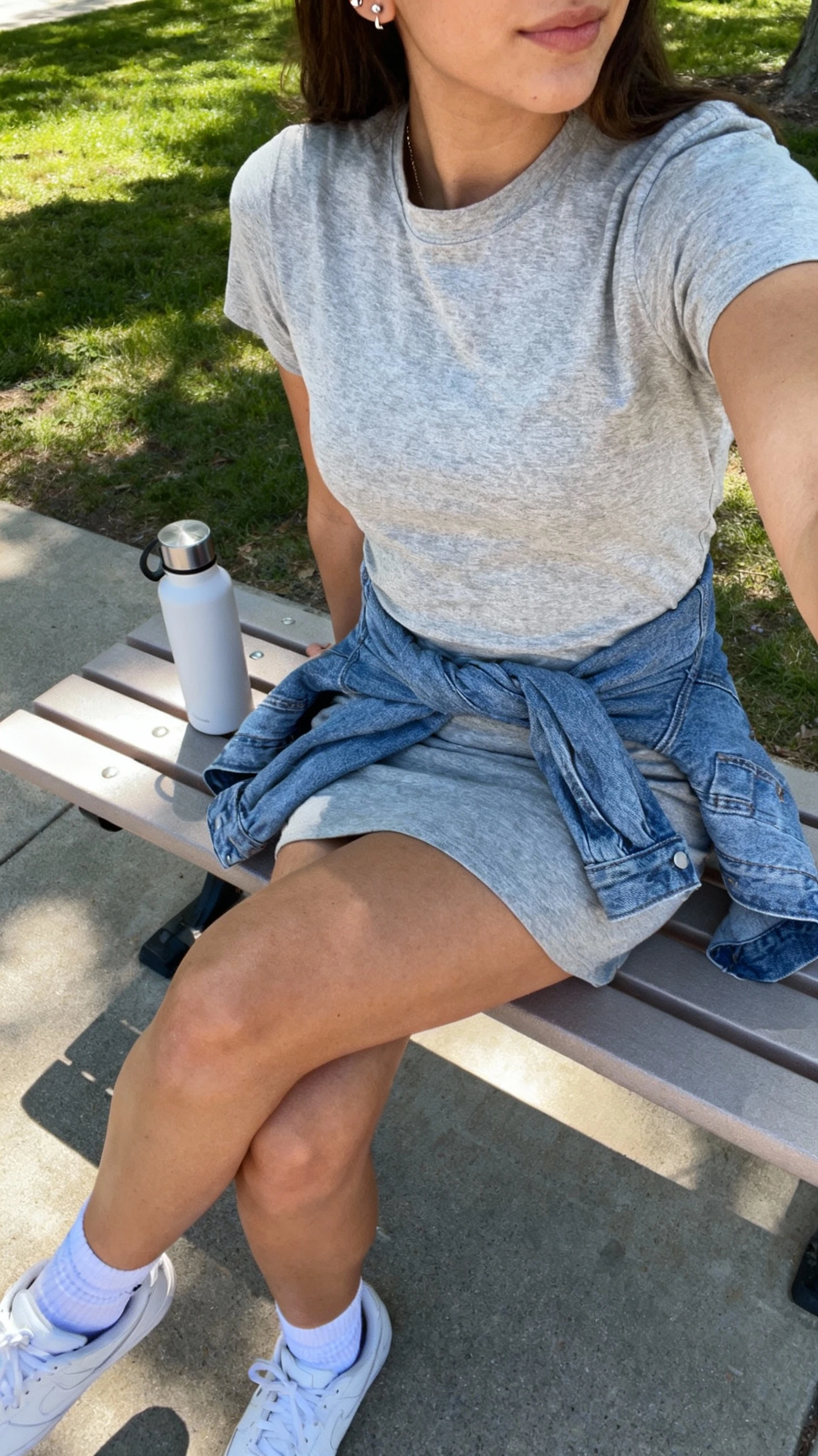 iPhone side-angle seated selfie of a woman in an everyday T-shirt dress (heather gray) with white sneakers, ankle socks, denim jacket tied around waist, simple studs, reusable water bottle beside her, face not visible, campus quad bench in daylight, natural iPhone photo quality.