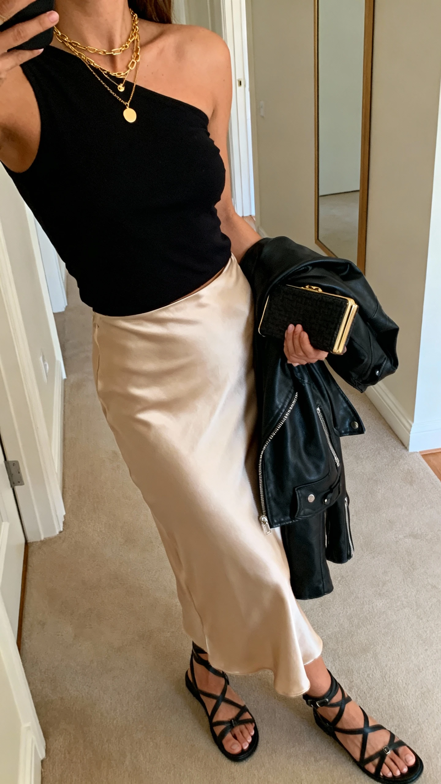 iPhone side-angle selfie cropped at shoulders of a woman wearing a black one-shoulder top with a champagne satin slip skirt, leather moto jacket draped over arm, strappy black sandals, layered gold necklaces, small clutch, face not visible, hallway mirror, warm indoor lighting, natural iPhone aesthetic.