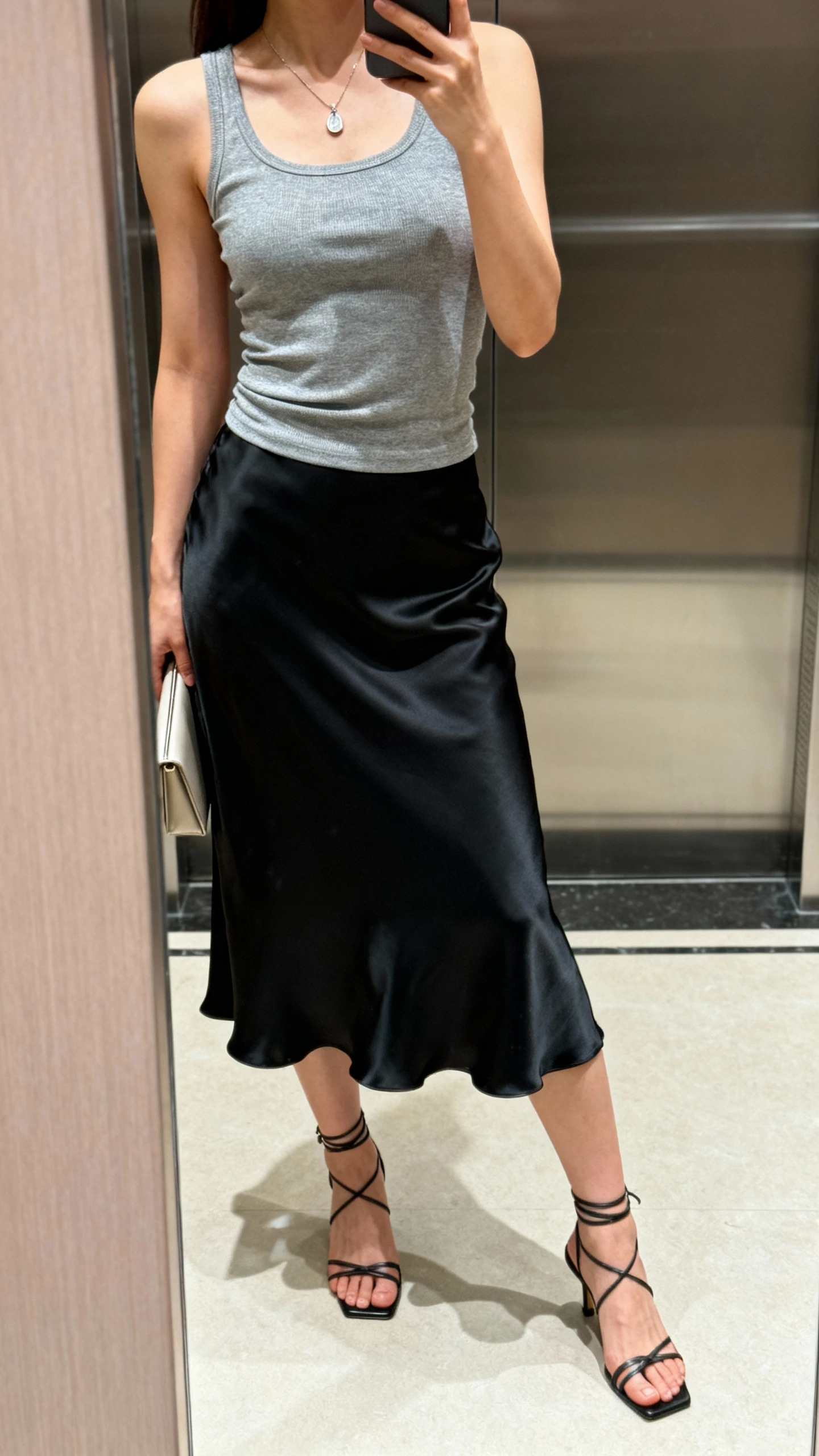 iPhone side-angle selfie of a woman in a black silk slip skirt, fitted gray tank, delicate pendant necklace, strappy heeled sandals, and a minimalist clutch; face not visible, elevator mirror, soft indoor lighting, natural iPhone photo.