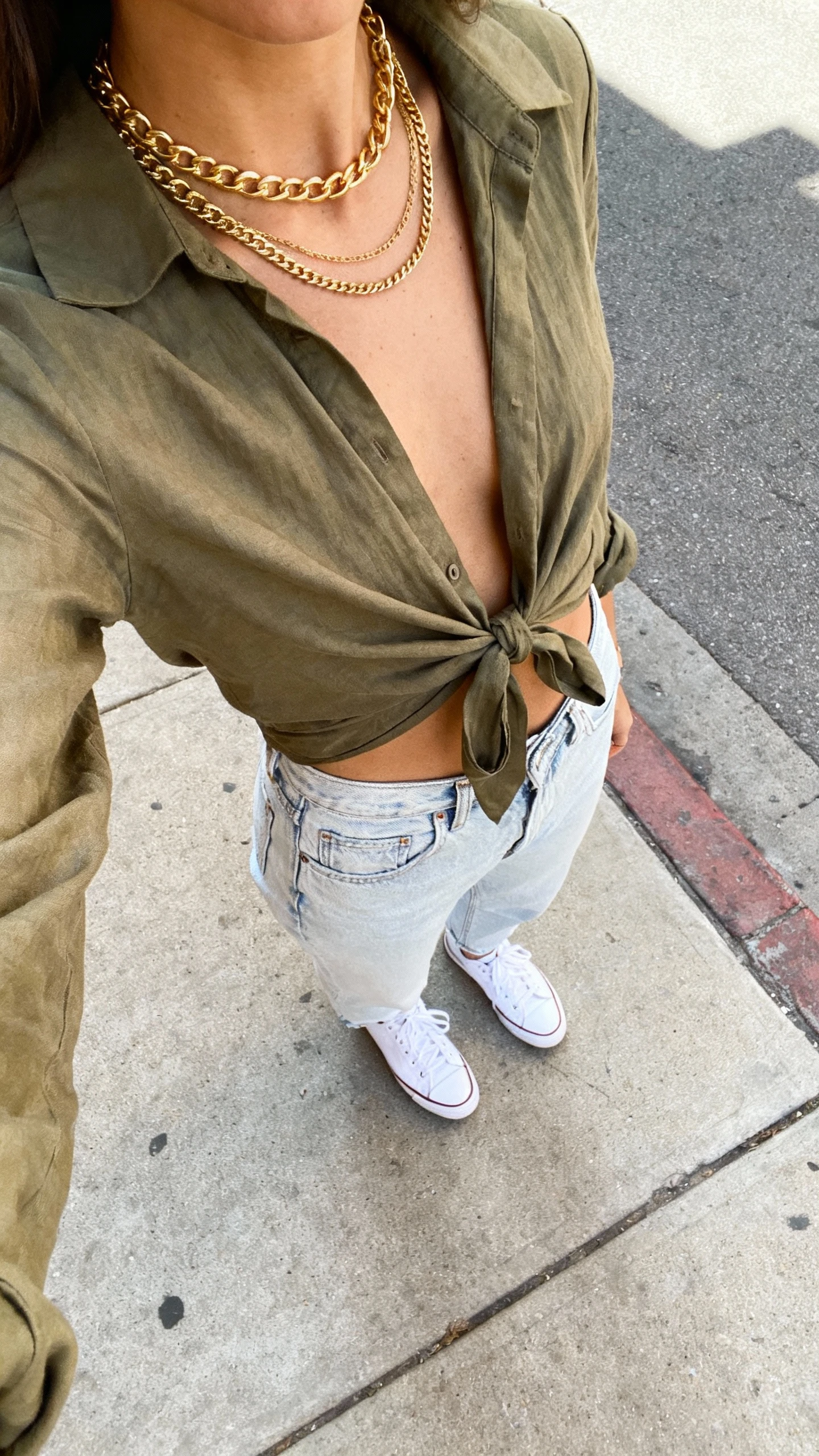 iPhone sidewalk selfie, downward angle, of a woman in a tie-front blouse in olive, light-wash mom jeans, white low-top sneakers, layered gold chains, face not visible, natural daylight on a city street, natural iPhone photo quality