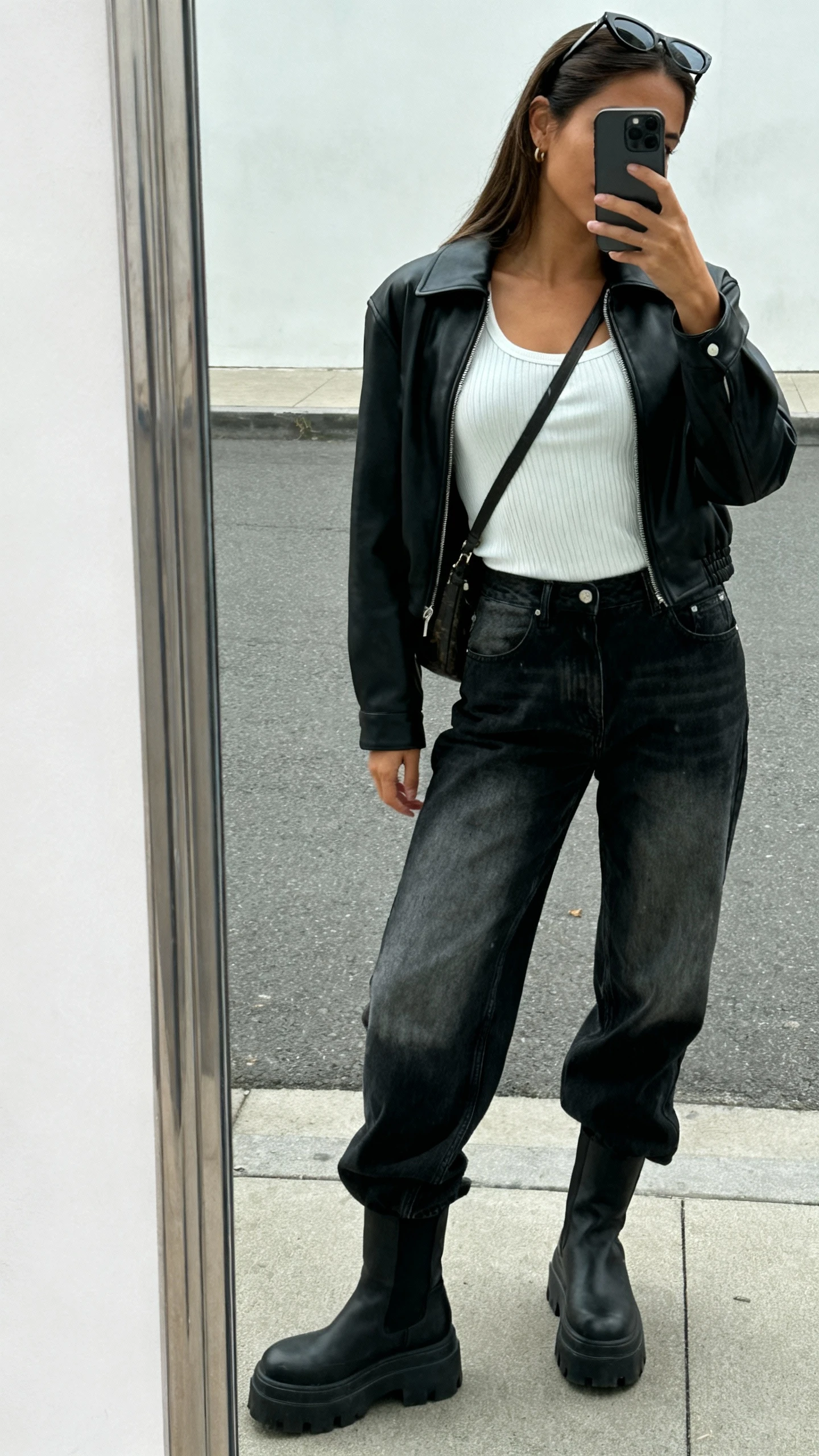 iPhone street-mirror selfie of a woman wearing a black leather jacket over a white ribbed tank, high-waist faded black jeans, chunky boots, slim sunglasses on head, and a crossbody; face not visible, side angle, overcast daylight, casual iPhone photo.