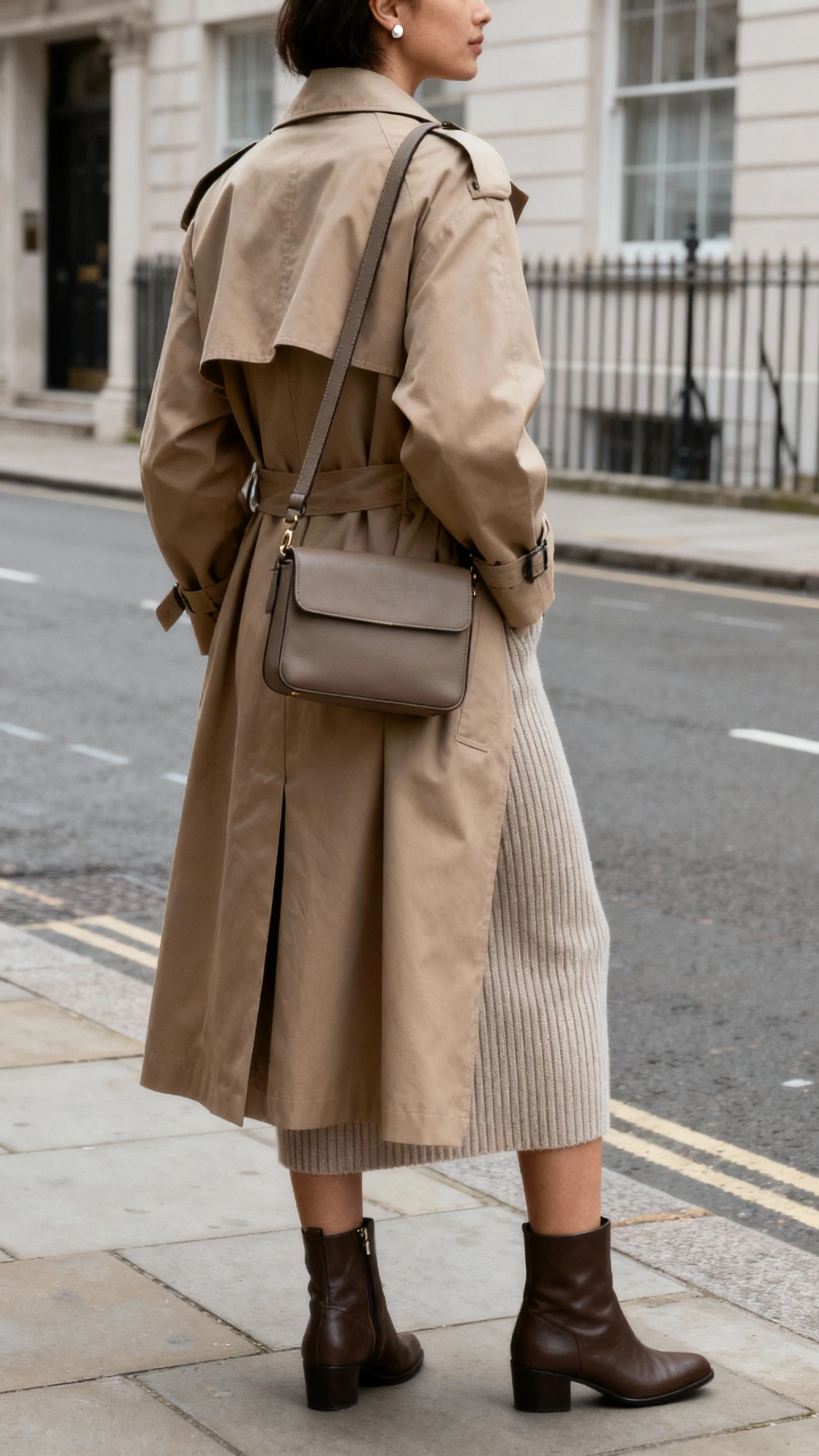 iPhone street-side selfie of a woman wearing a trench coat over a ribbed knit dress, ankle boots, structured crossbody, simple studs; face not visible, back/three-quarter view, overcast daylight on city sidewalk, natural iPhone quality.
