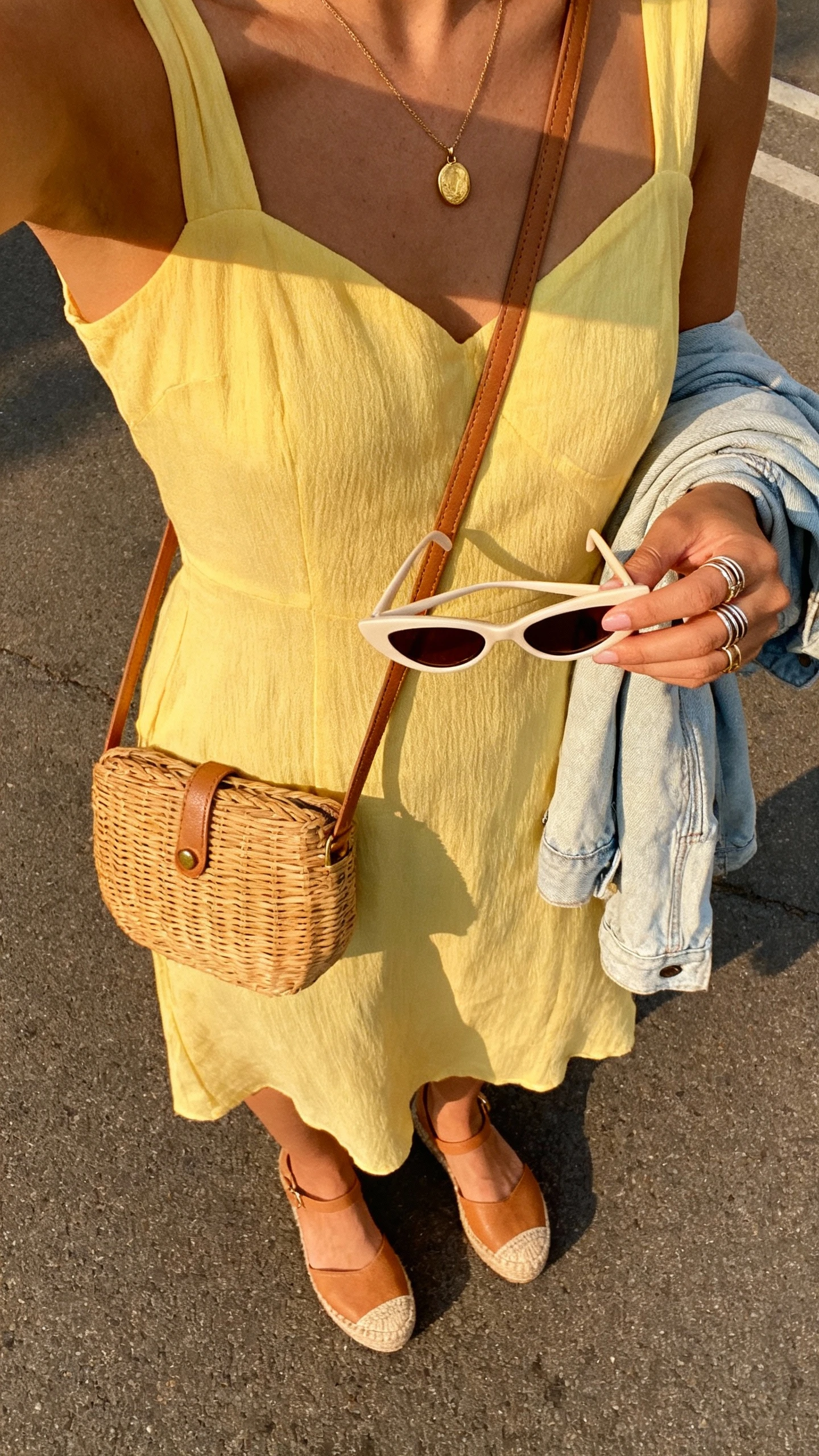 iPhone street-style selfie from chest down of a woman in a butter yellow sundress with tan espadrille wedges, wicker crossbody, cat-eye sunglasses in hand, simple gold pendant and stackable rings, light denim jacket draped over arm, face not visible, golden hour daylight, natural iPhone look.