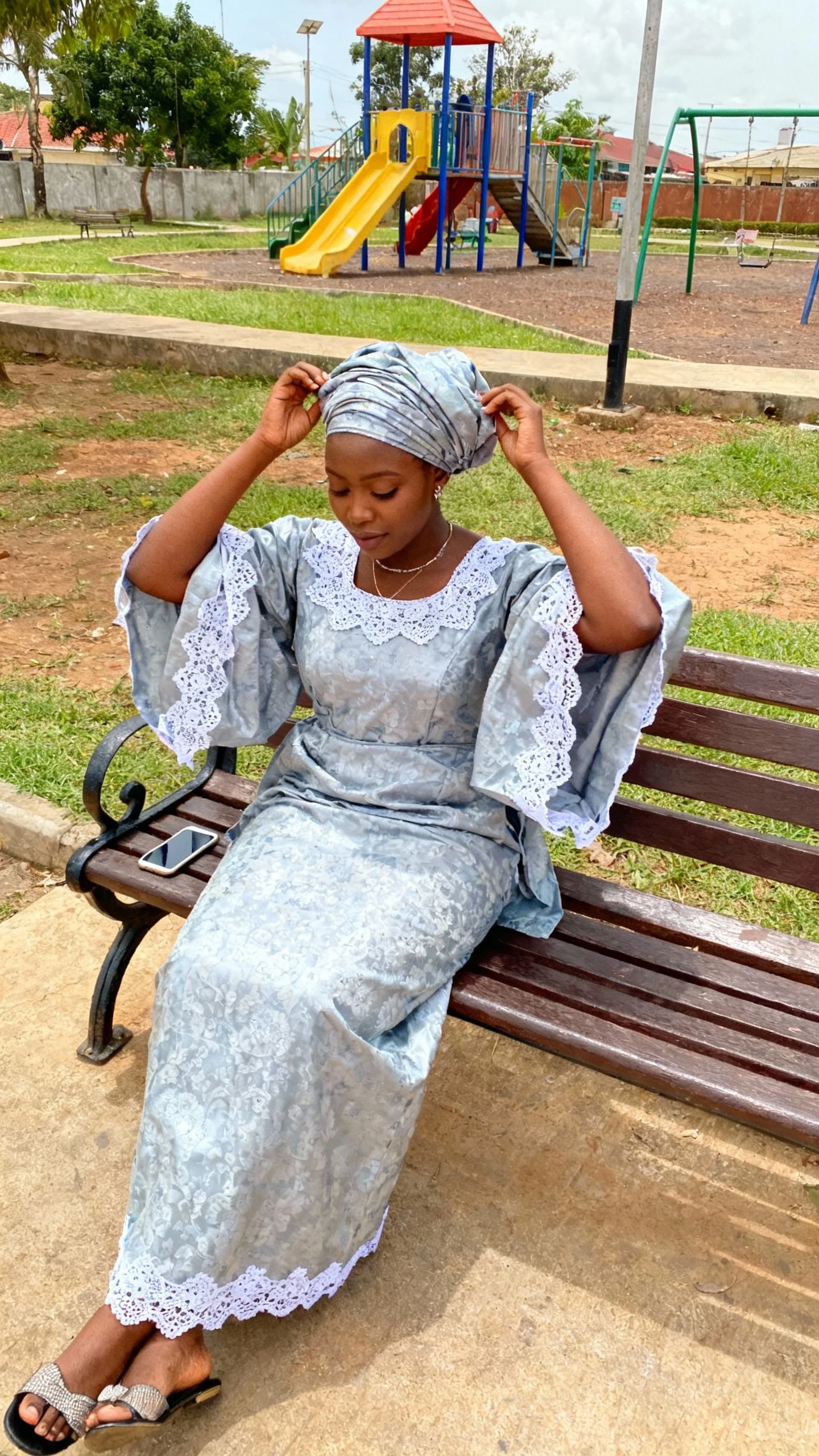 A woman wearing a classic iro and buba with lace accents, adjusting her headscarf on a bench near a community park, casual iPhone photo style, natural daylight, outdoor setting.