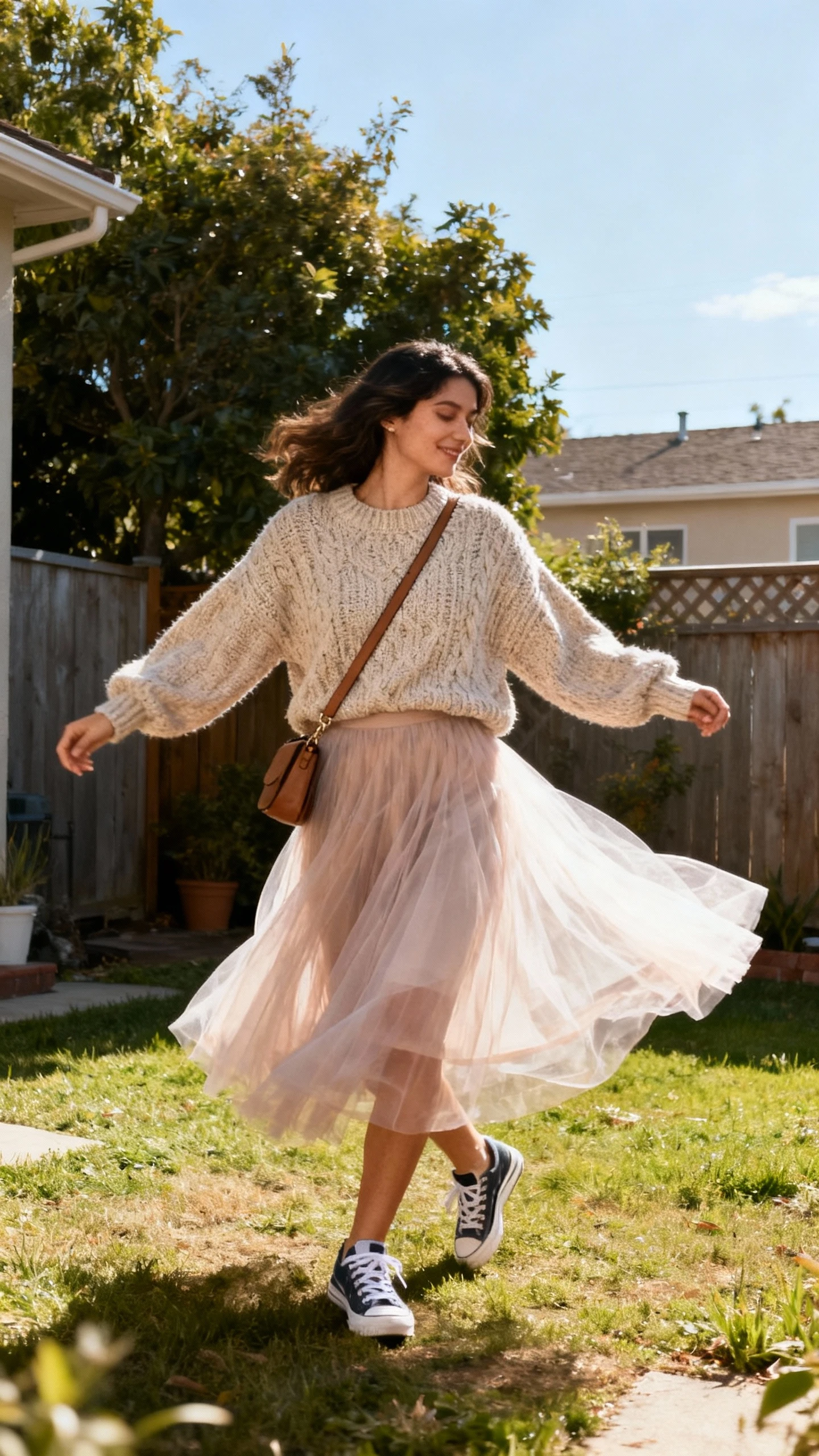A woman wearing a floaty tulle midi skirt paired with a chunky knit sweater, sneakers, and a crossbody bag, spinning in a backyard, candid moment, casual iPhone photo style, sunny day, outdoor setting.