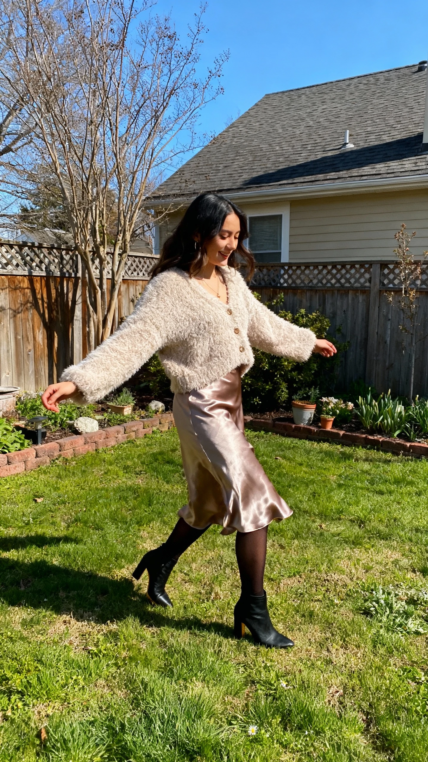 A woman wearing a fuzzy cardigan with a satin slip skirt, tights, and heeled booties, twirling slightly by a backyard garden, casual iPhone photo style, natural daylight, outdoor setting.