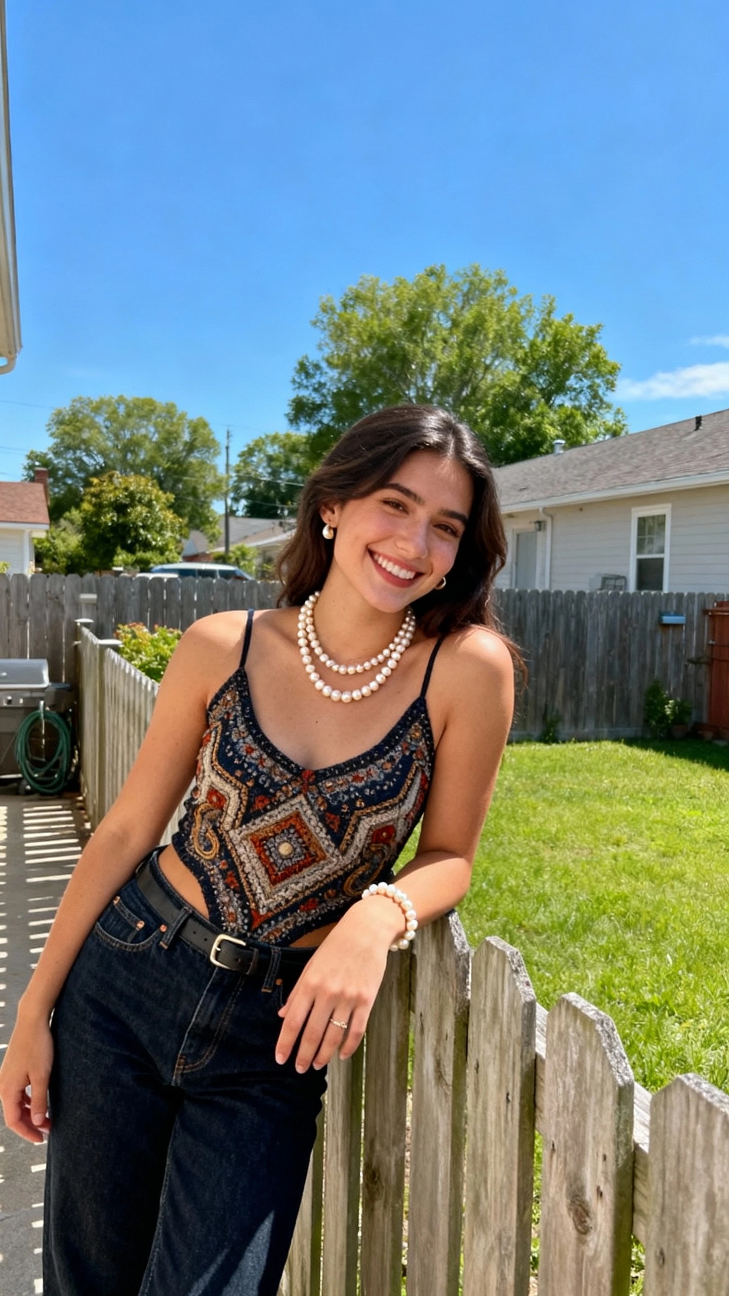 A woman wearing dark denim, a statement top, and pearly accessories, leaning on a backyard fence with a relaxed grin, casual iPhone photo style, sunny day, outdoor setting.