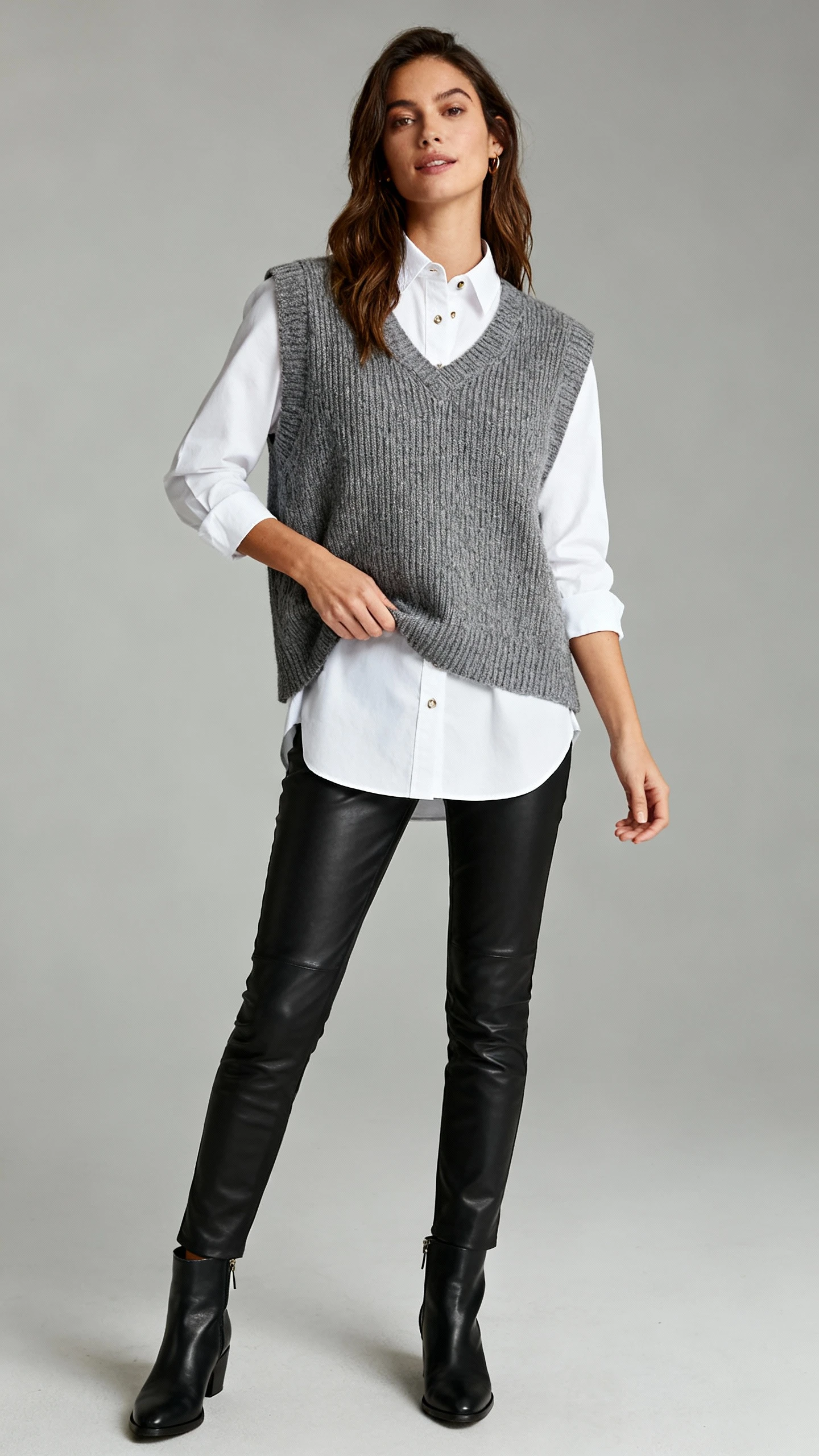 Editorial studio photo of a woman layering a sleeveless knit over a crisp white button-down with slim leather pants and sleek ankle boots, neutral gray background, soft lighting, natural stance, clean composition.