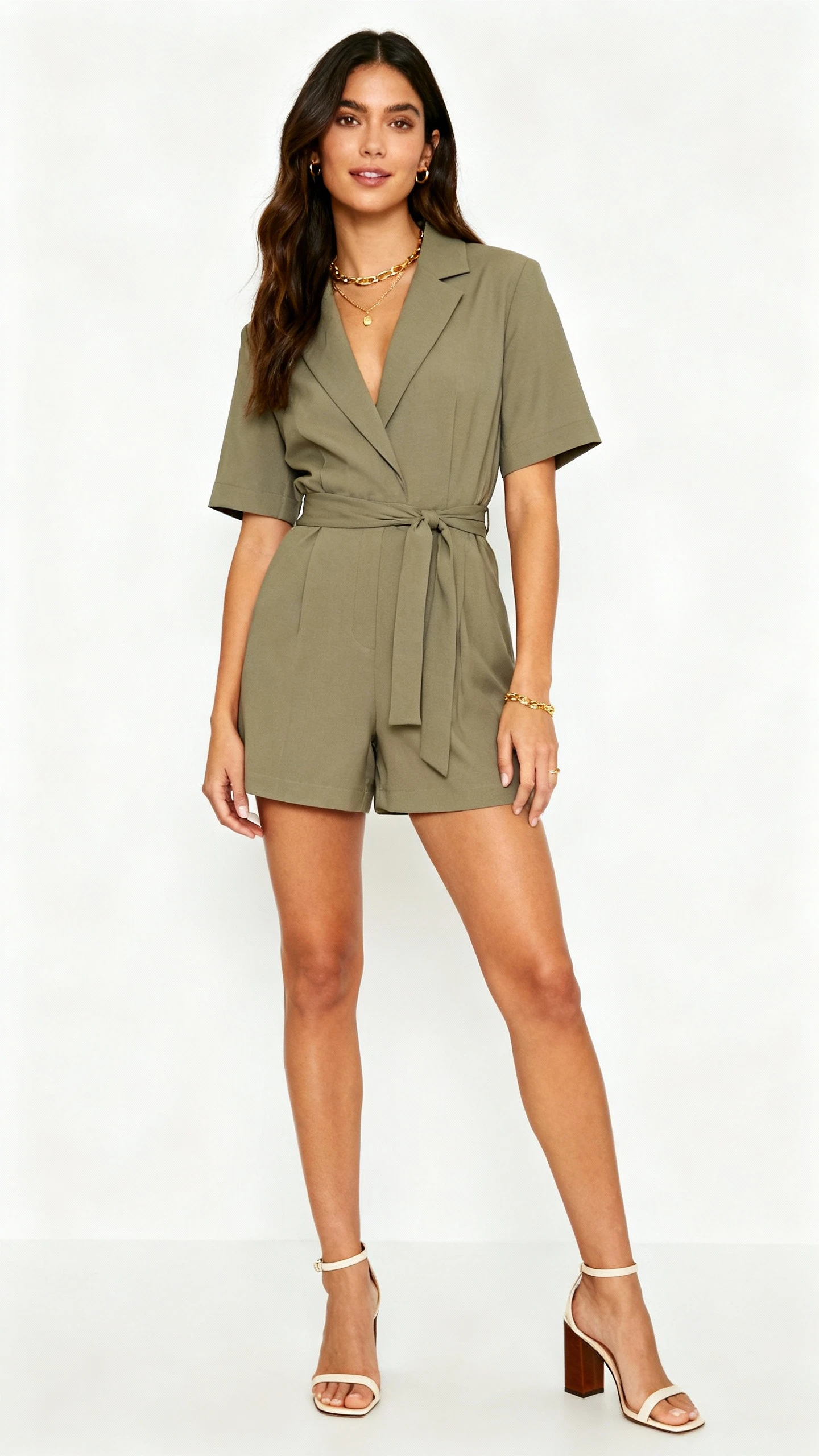 Editorial style studio photo of a woman wearing an elevated tailored romper with a cinched waist, block-heel sandals, and subtle gold jewelry, white background, soft lighting, professional natural pose, clean and polished.