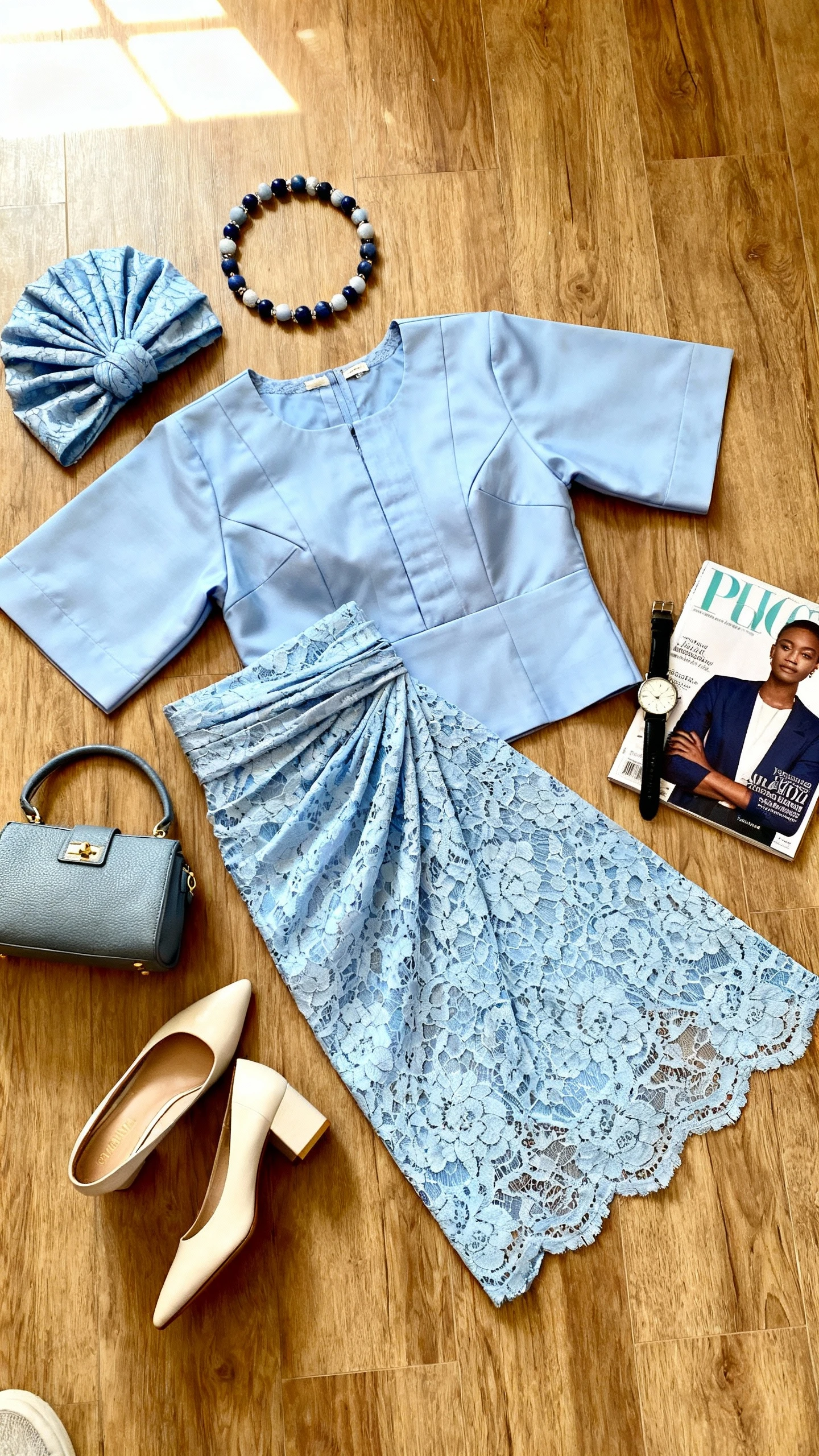 Flat lay overhead of a modern lace iro (wrapped skirt) and tailored buba (top) set with matching head tie, low block heels, beaded necklace, and a structured handbag on a wooden floor; add a watch and a fashion magazine, bright natural light, no person visible, organized composition, iPhone photo aesthetic.