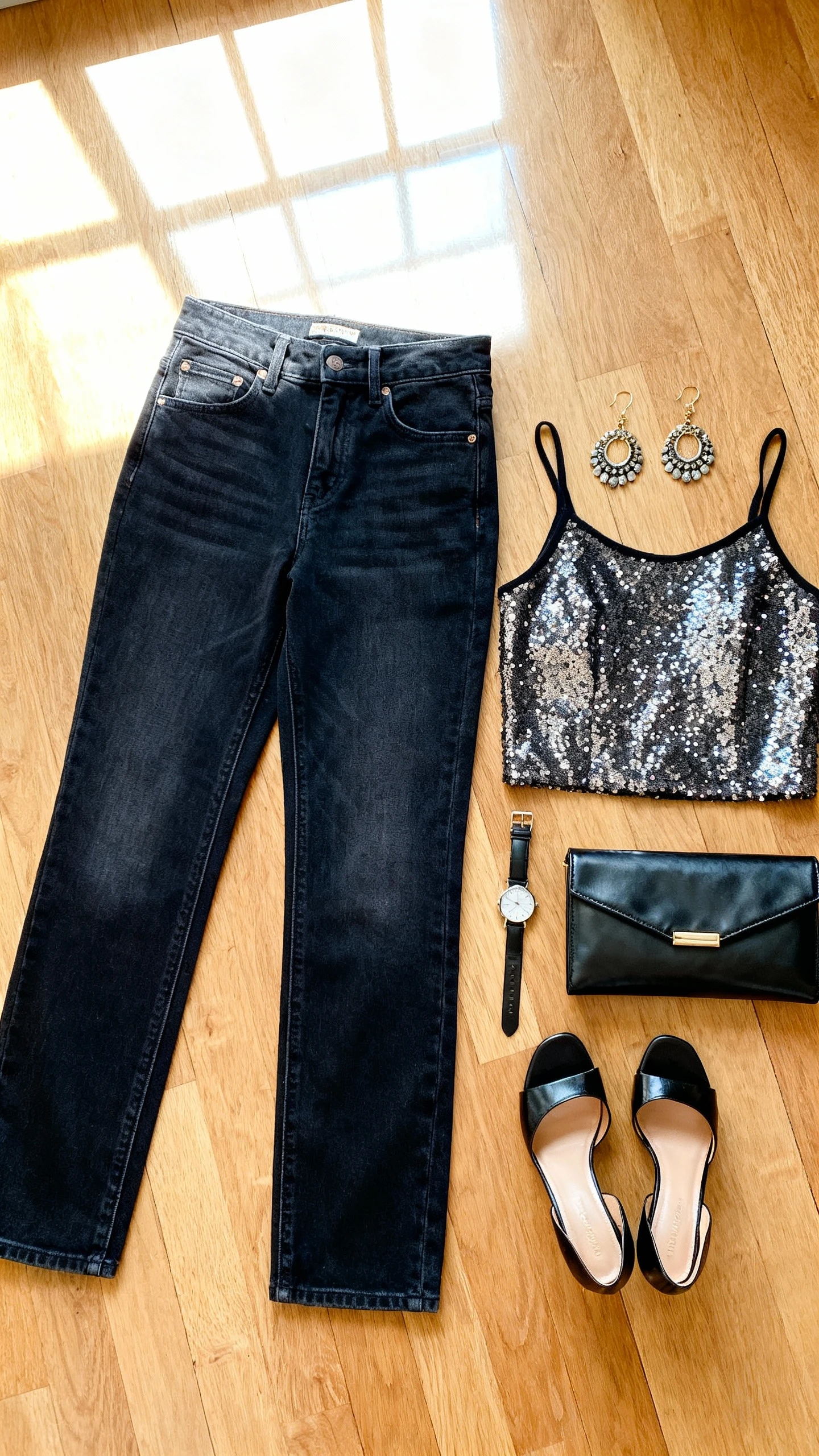 Flat lay overhead of high-rise dark-wash jeans, sparkly party top, heeled mules, statement earrings, and a sleek clutch on wooden floor; include a watch as prop, bright natural window light, no person visible, organized composition, iPhone photo aesthetic.
