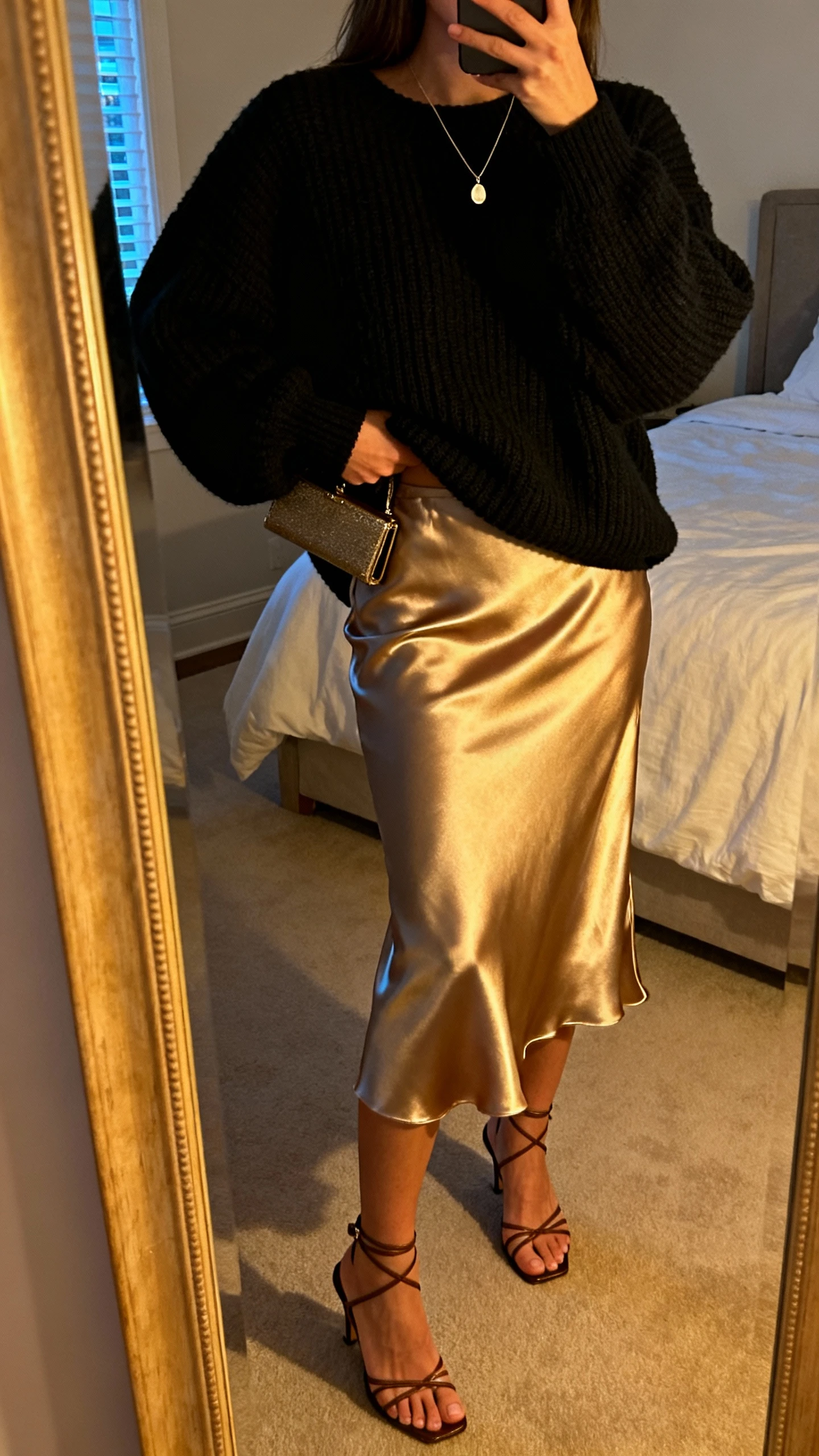 iPhone back-view mirror selfie of a woman wearing a slinky satin slip skirt with an oversized black knit sweater slightly tucked at the front, strappy heeled sandals, a delicate pendant necklace, and a small clutch in hand, face not visible, bedroom mirror, warm evening indoor light, iPhone photo quality.