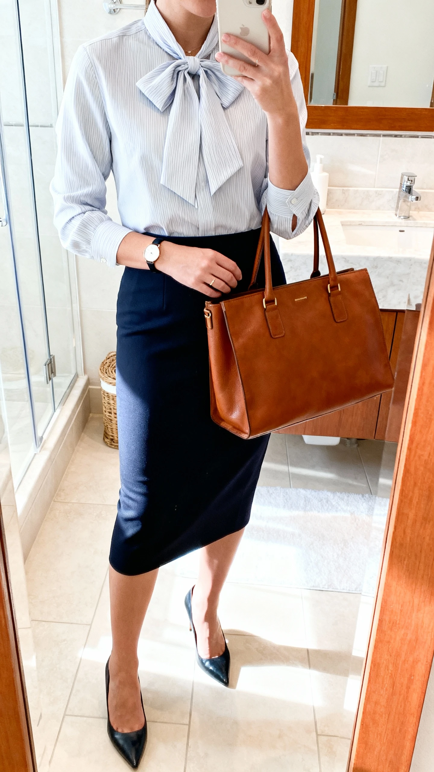 iPhone mirror selfie of a woman in a bow-tie blouse tucked into a pencil skirt with classic pumps, leather tote and a slim watch for office-to-dinner polish, face not visible, cropped at neck, bathroom mirror, bright indoor light, iPhone photo quality.