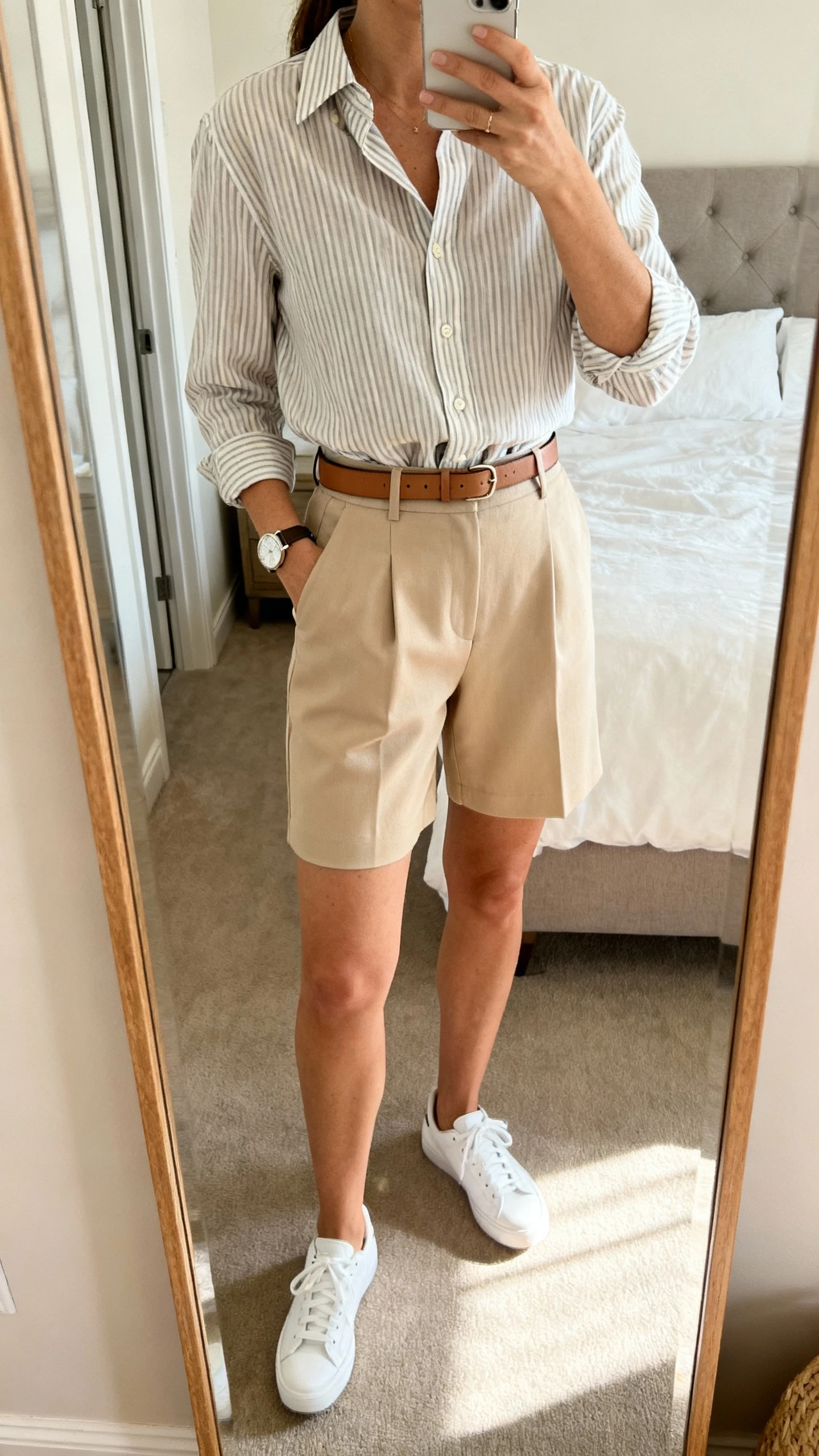 iPhone mirror selfie of a woman in a lightweight striped button-down half-tucked into tailored beige shorts, with a slim leather belt, white sneakers, and a simple watch, face not visible, cropped at shoulders, bedroom mirror, mid-morning light, iPhone photo quality.