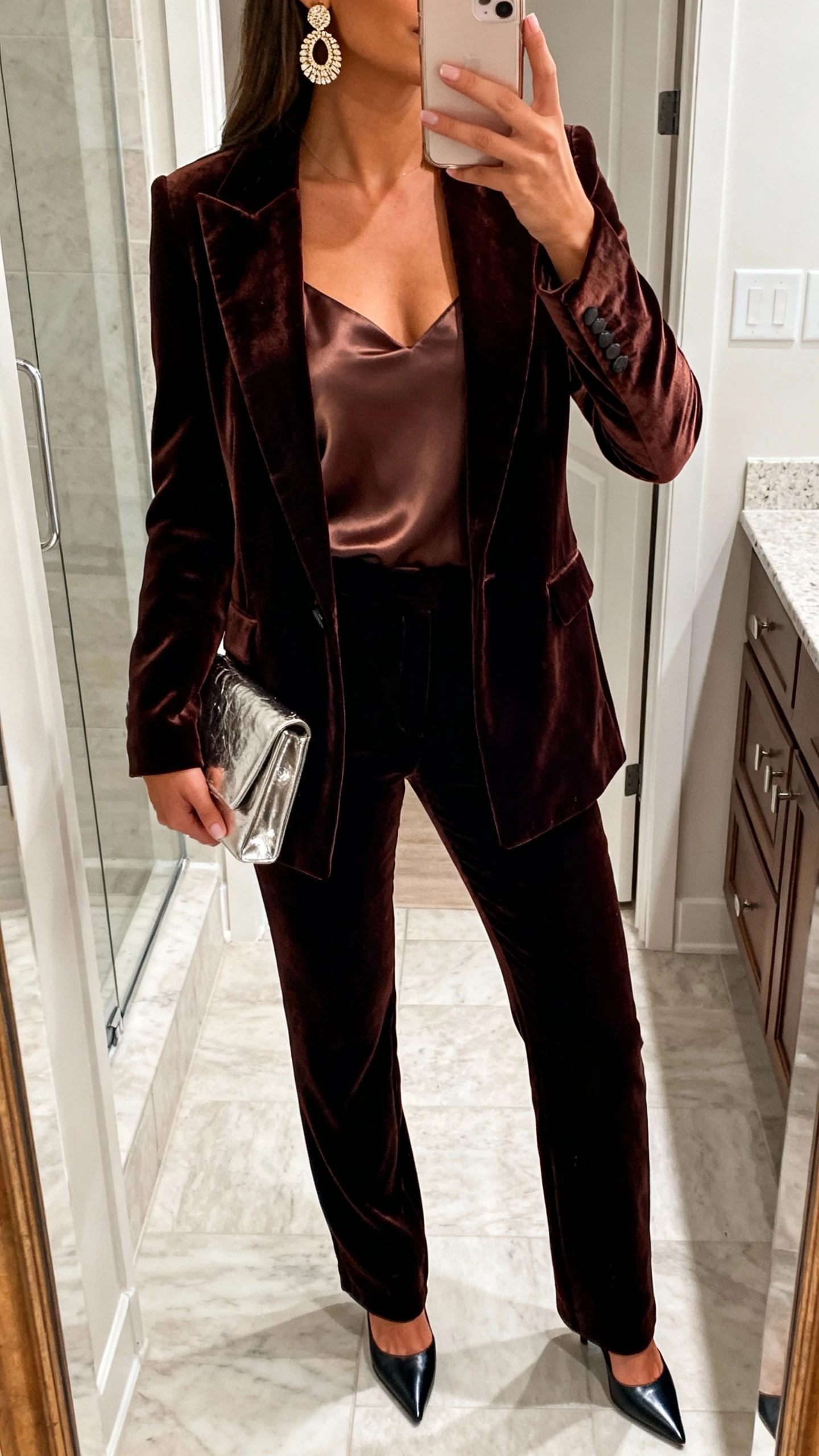 iPhone mirror selfie of a woman in a night-out velvet power suit: tailored velvet blazer and matching trousers, silky camisole, pointed-toe heels, statement earrings, and a metallic clutch; face not visible, cropped at neck, bathroom mirror, soft indoor lighting, iPhone photo quality.