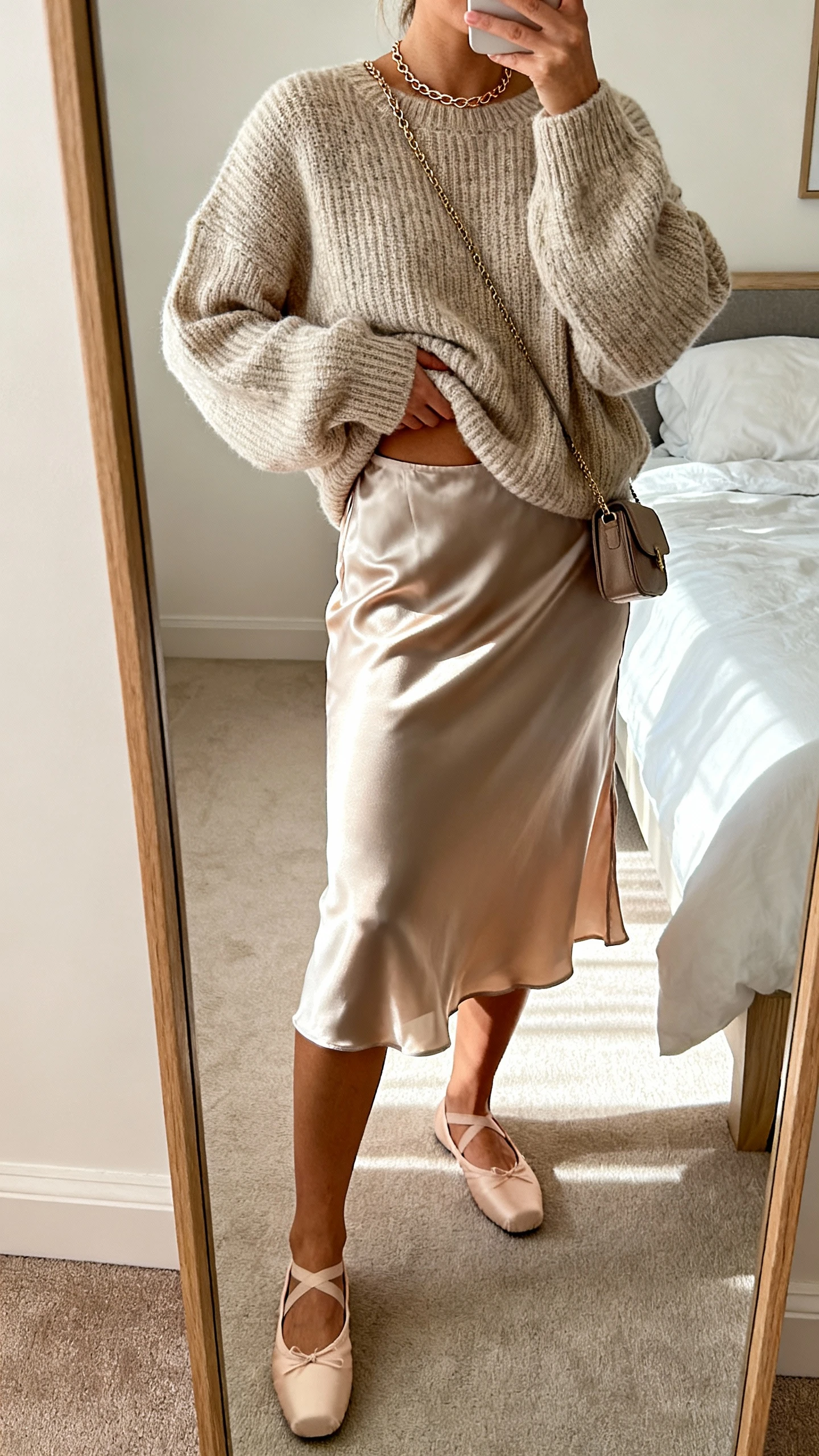 iPhone mirror selfie of a woman in a satin slip skirt with a cozy oversized knit sweater half-tucked, delicate chain, ballet flats, and a small crossbody, face not visible, cropped at shoulders, bedroom mirror, soft morning light, iPhone photo quality.