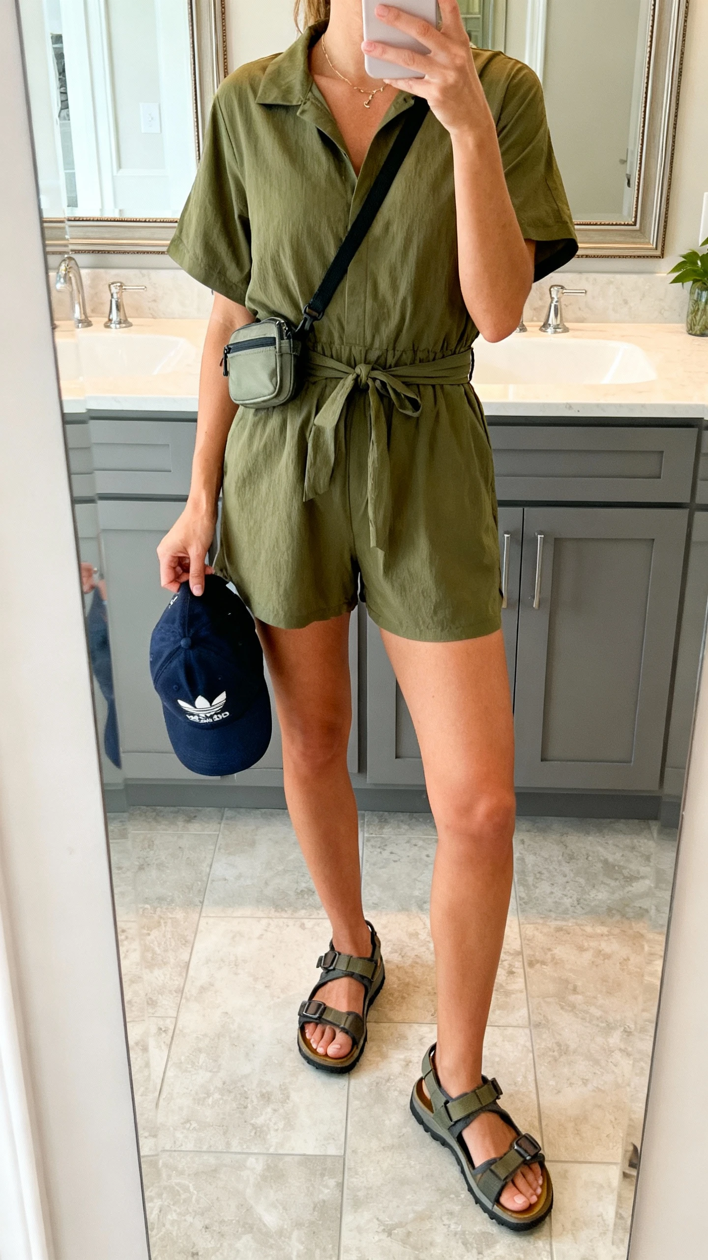 iPhone mirror selfie of a woman in a sporty-chic olive romper with a cinched waist, trail sandals, a baseball cap in hand, and a small crossbody belt bag, face not visible, cropped at neck, bathroom mirror, natural indoor light, iPhone photo quality.
