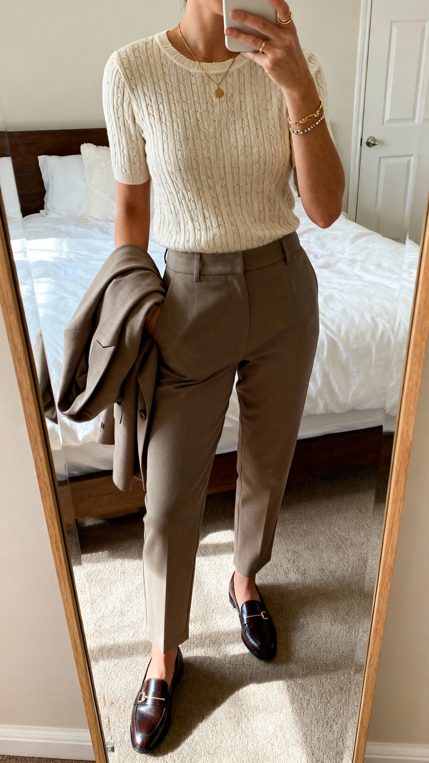 iPhone mirror selfie of a woman in a week-ready work look: refined knit top, tailored pants, blazer over arm, classic loafers, and understated jewelry, face not visible, cropped at neck, bedroom mirror, natural morning light, iPhone photo quality